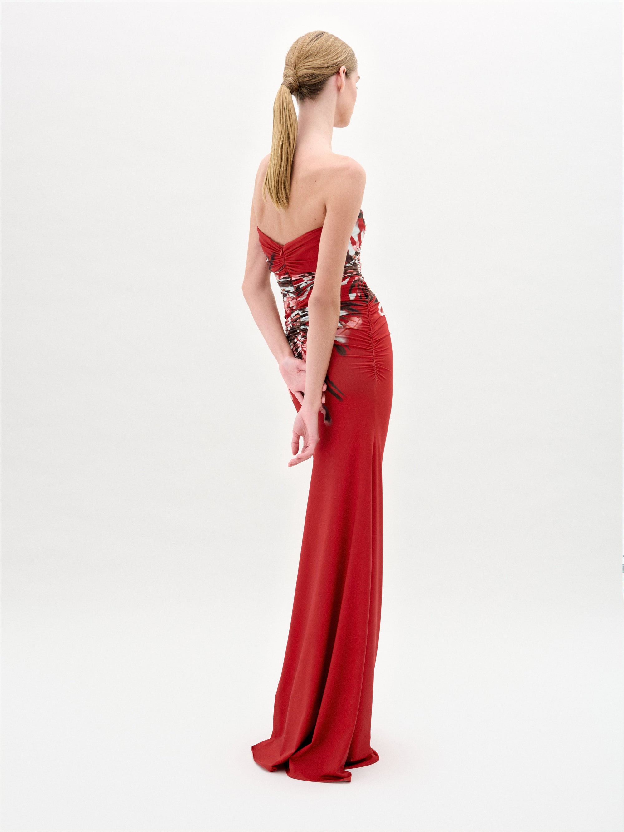 A woman with blonde hair in a ponytail wears the Collins Dress, a strapless, form-fitting, floral-patterned red gown by SVT DESIGNS. She stands with her back to the camera against a plain white background.