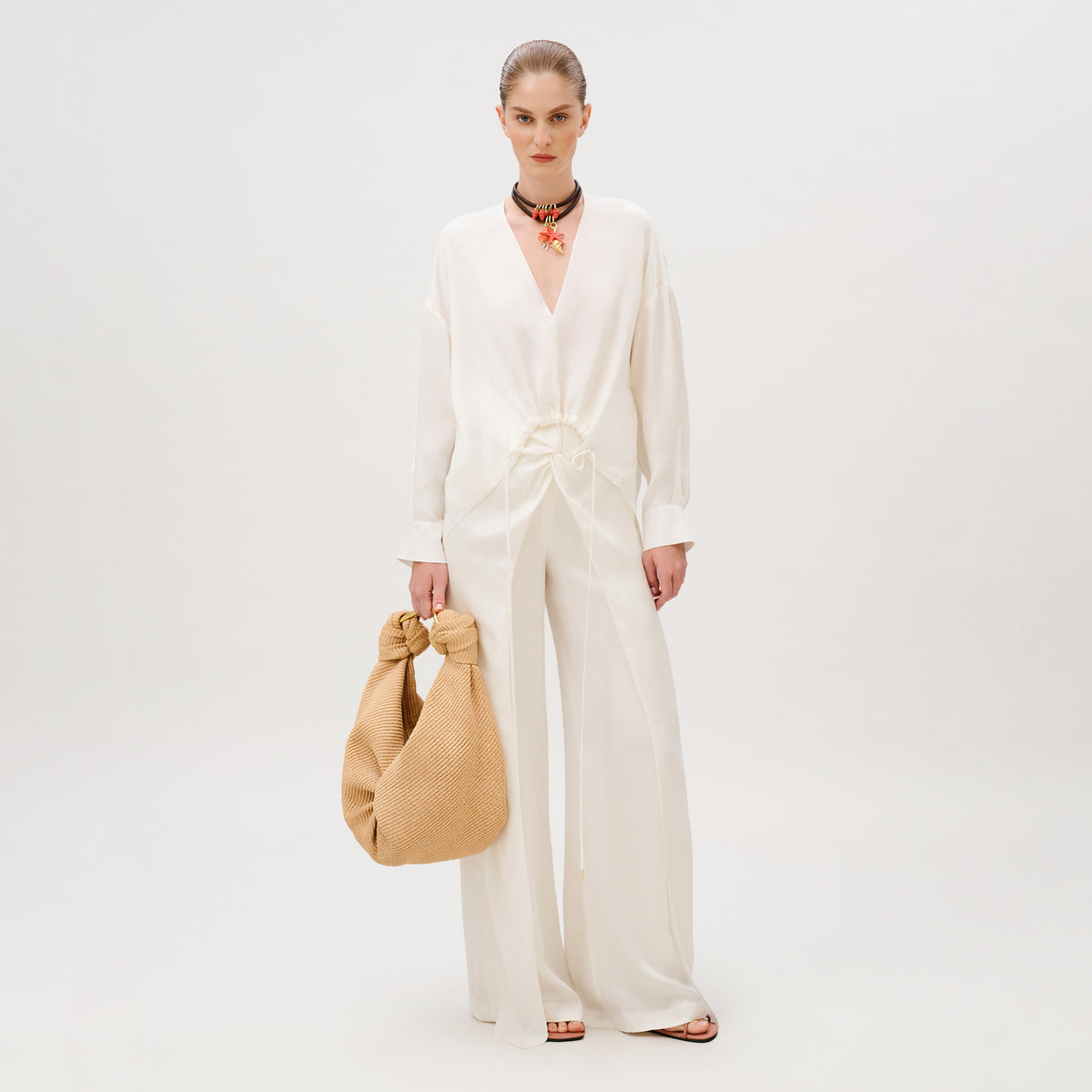A model wearing a white long-sleeve v-neck blouse and wide-leg Canturipe Pant, carrying a large beige knotted handbag.