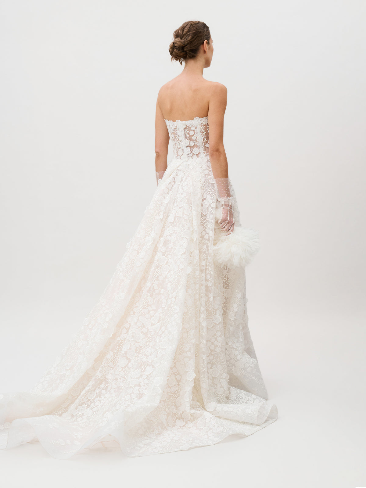 A woman faces away, holding a white, fluffy clutch while wearing the Cindy Dress—a strapless lace wedding gown expertly crafted from luxurious Italian fabric by a renowned Colombian atelier. The dress showcases a long, elegant train and sheer gloves against a plain white background.