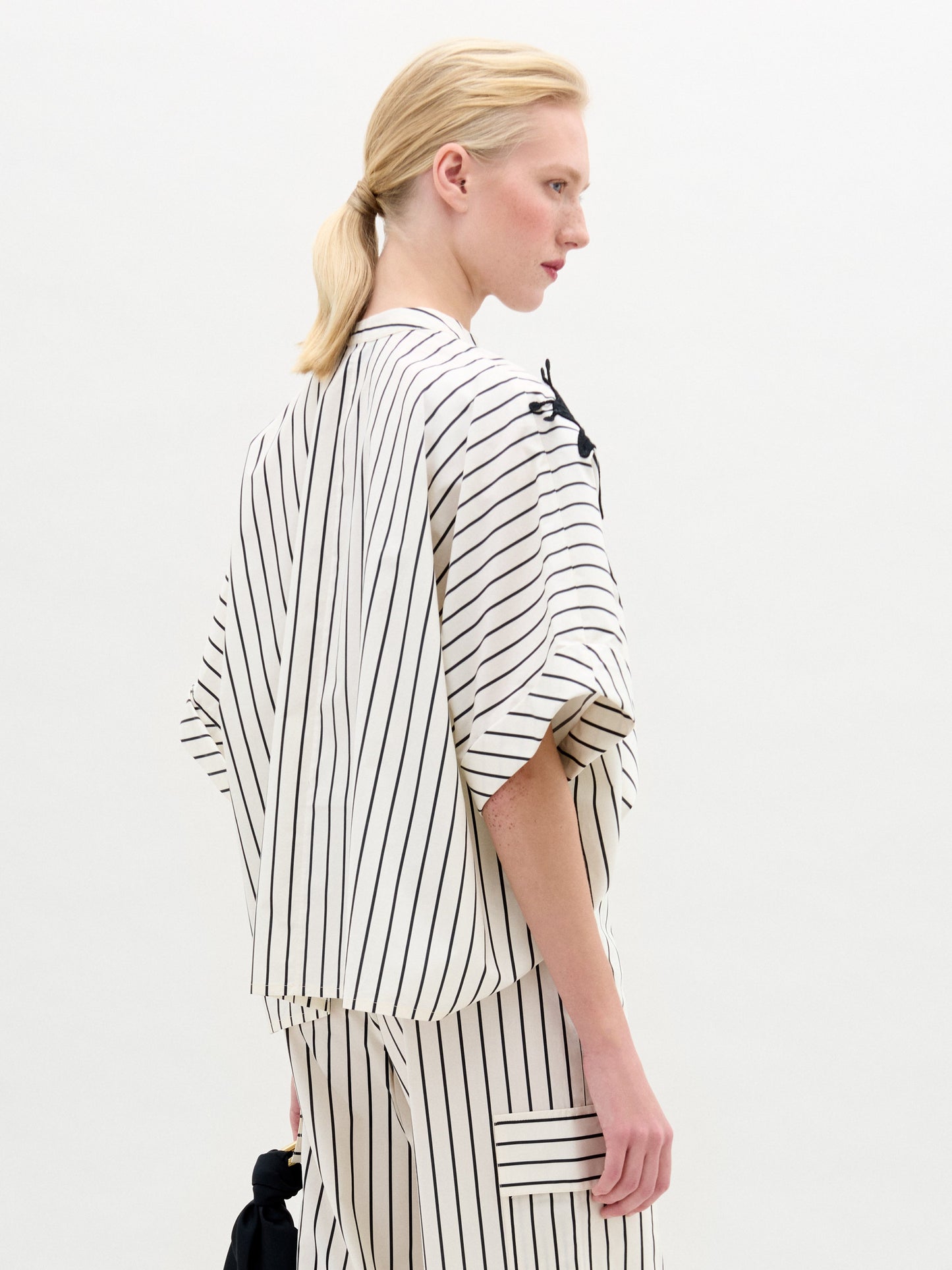 A person with blonde hair in a low ponytail wears the Daia Blouse by SVT DESIGNS, a loose, cream cotton top with vertical black stripes, wide sleeves, and pocket detail, standing against a plain light background.