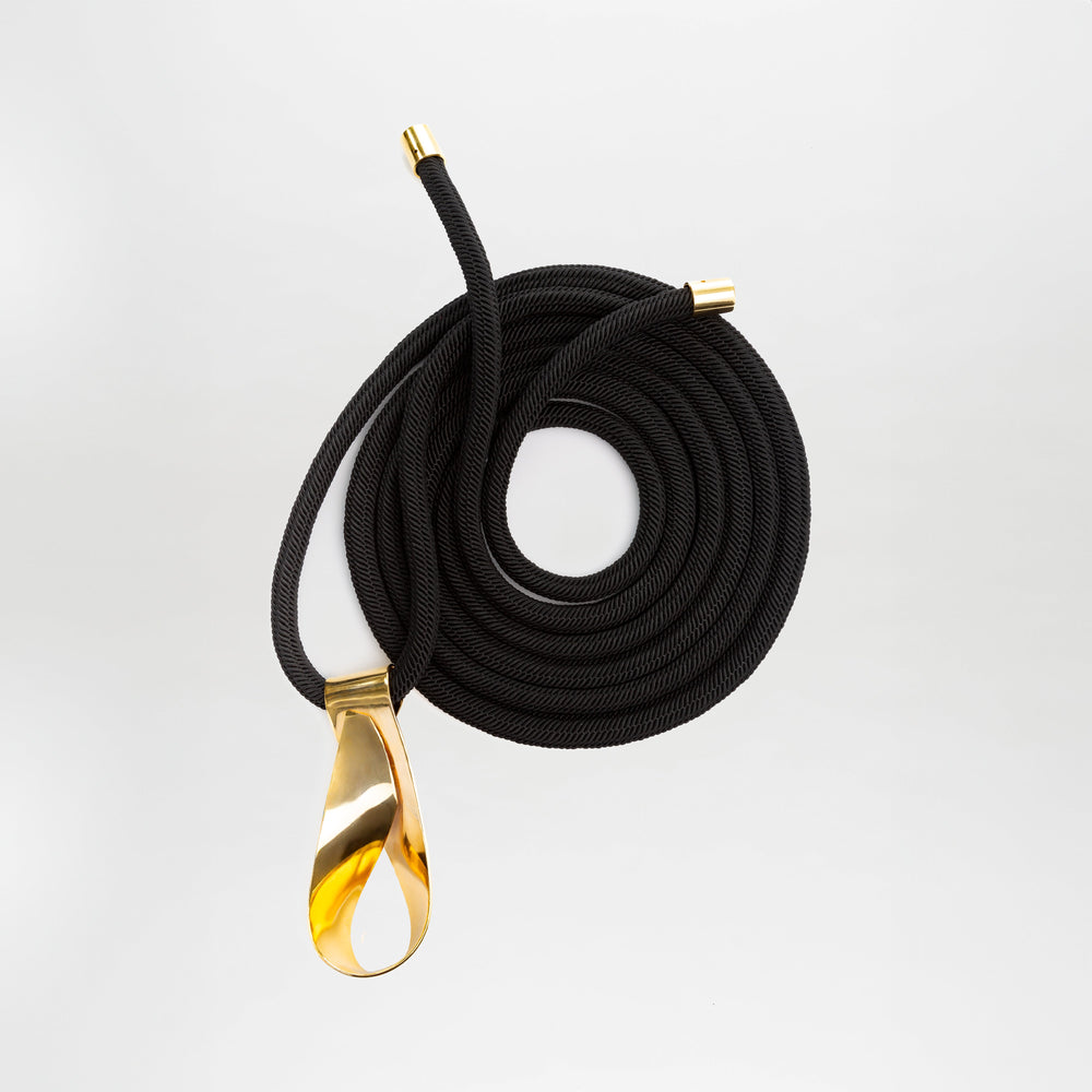 The Daiana Belt by SVT DESIGNS features a coiled black cord with gold metal caps and a large teardrop-shaped, gold-plated brass buckle, shown against a plain white background.