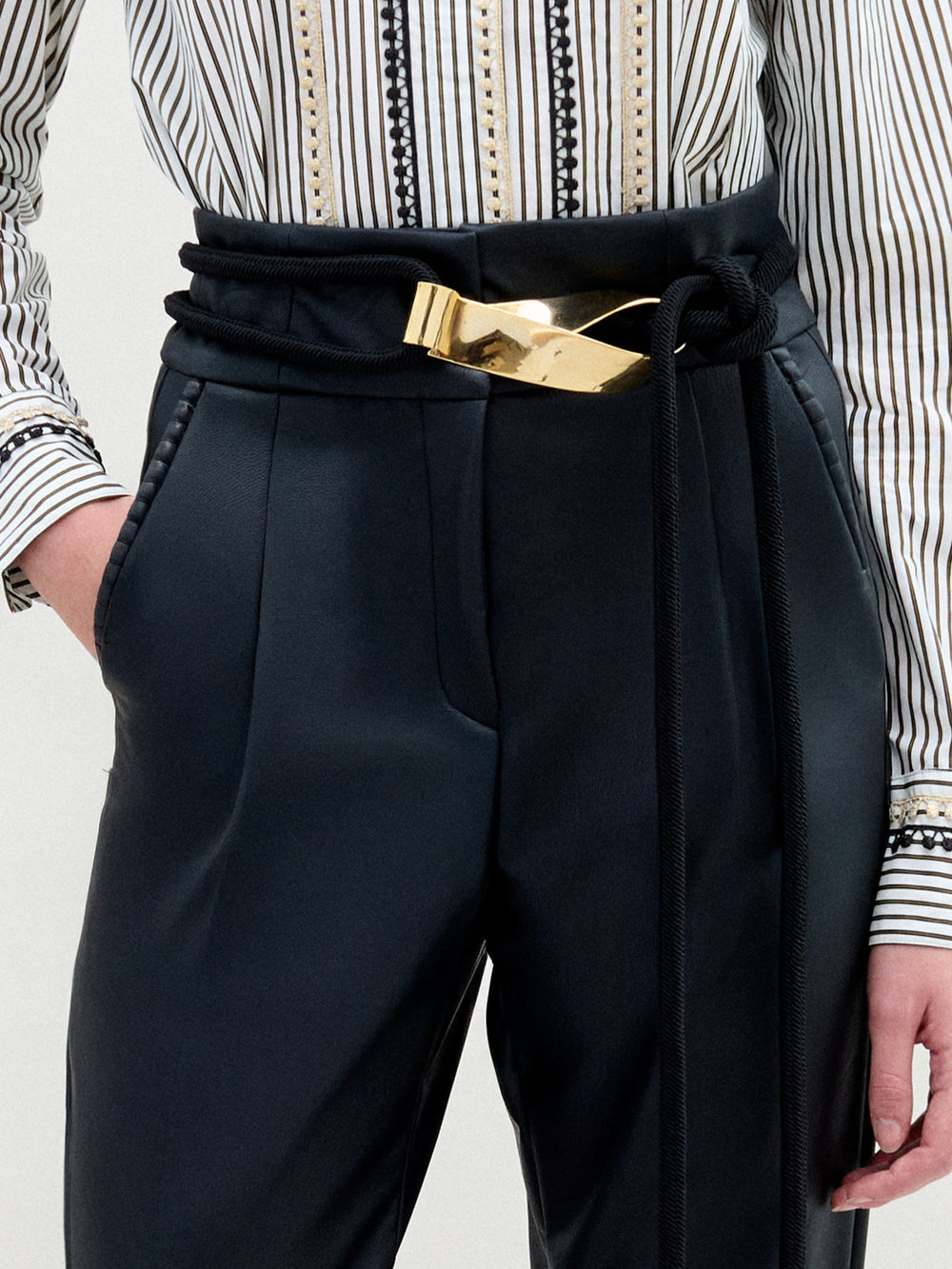 A person wears black high-waisted trousers styled with the SVT DESIGNS Daiana Belt, featuring a gold-plated brass buckle, and a striped shirt with decorative buttons and patterns; one hand rests in their pocket.