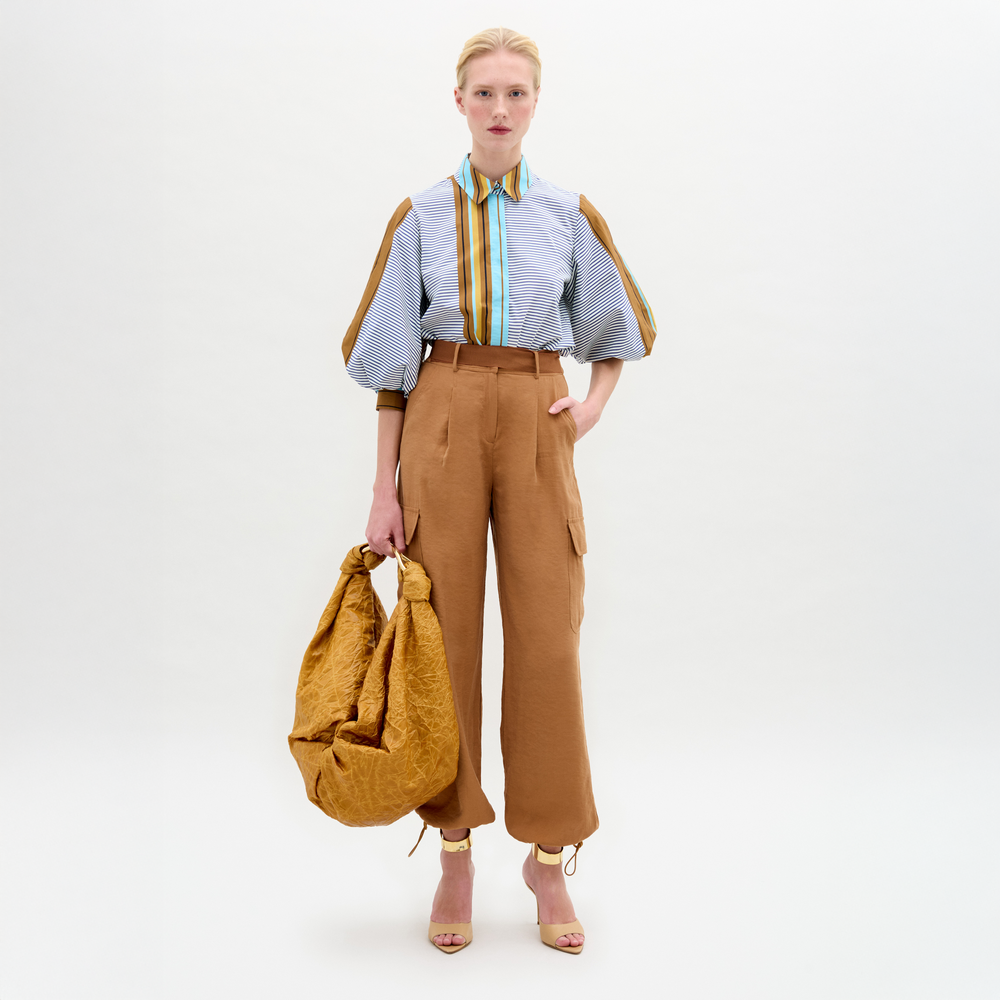 A person stands against a plain white background wearing a striped shirt, heeled sandals, holding a large brown textured bag, and relaxed fit high-waisted Darnell Pant by SVT DESIGNS in a cargo style.