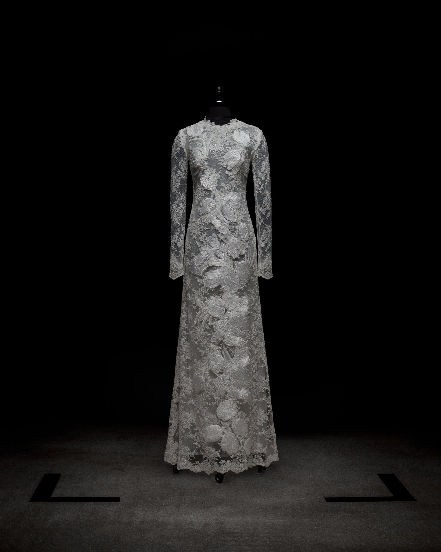 The Dava Dress by SVT DESIGNS is a full-length, long-sleeved gown with intricate lace patterns and floral designs from the high neckline to the hem, showcased on a black mannequin against a dark background.