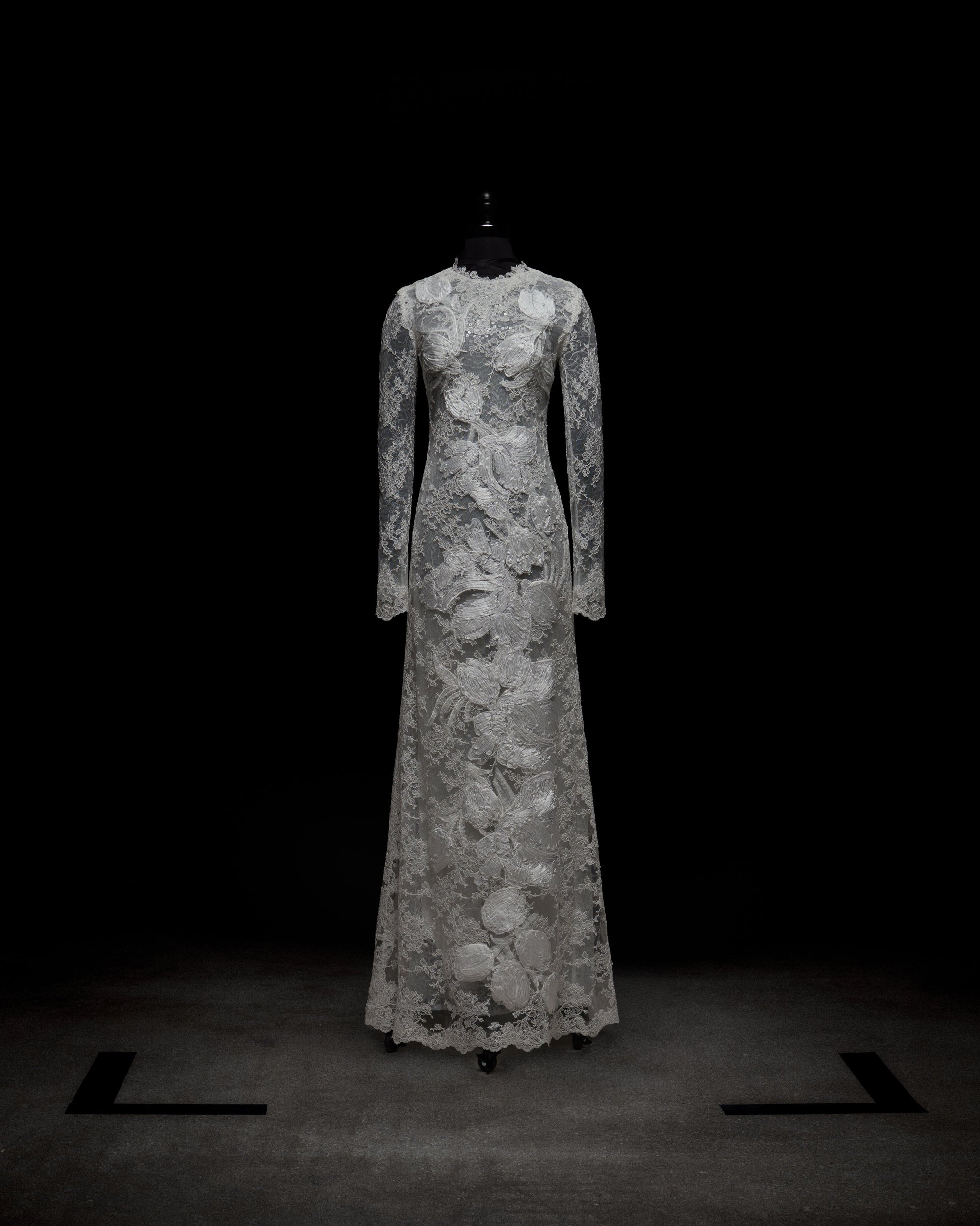 The Dava Dress by SVT DESIGNS is a full-length, long-sleeved gown with intricate lace patterns and floral designs from the high neckline to the hem, showcased on a black mannequin against a dark background.