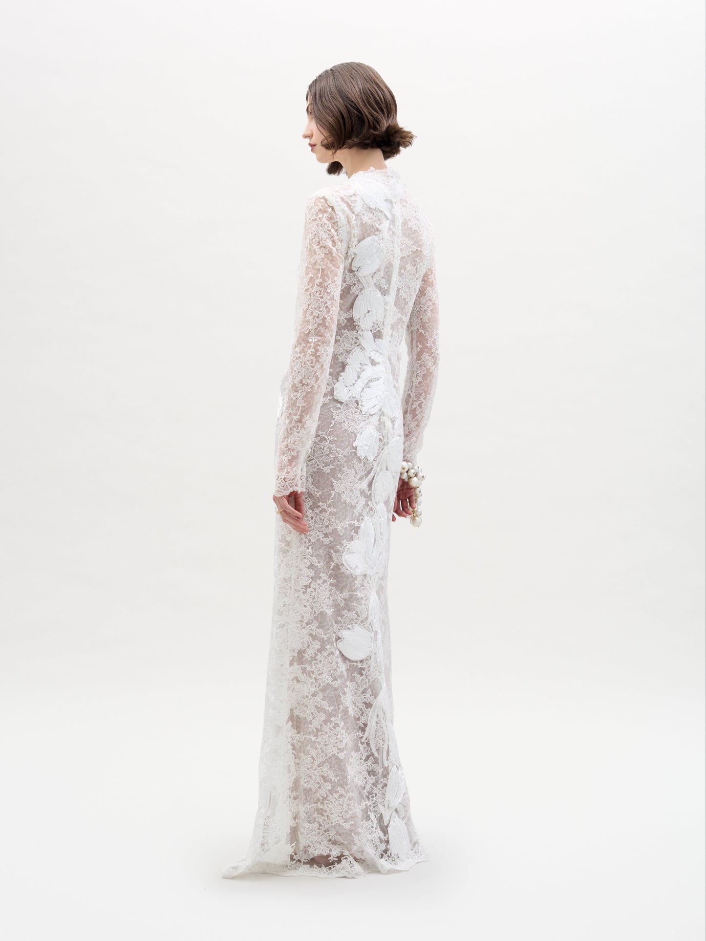A woman stands against a plain background wearing the SVT DESIGNS Dava Dress, showcasing its long-sleeved, floor-length design. The dress is white with a beautiful floral lace pattern as she faces away, highlighting the back and side.