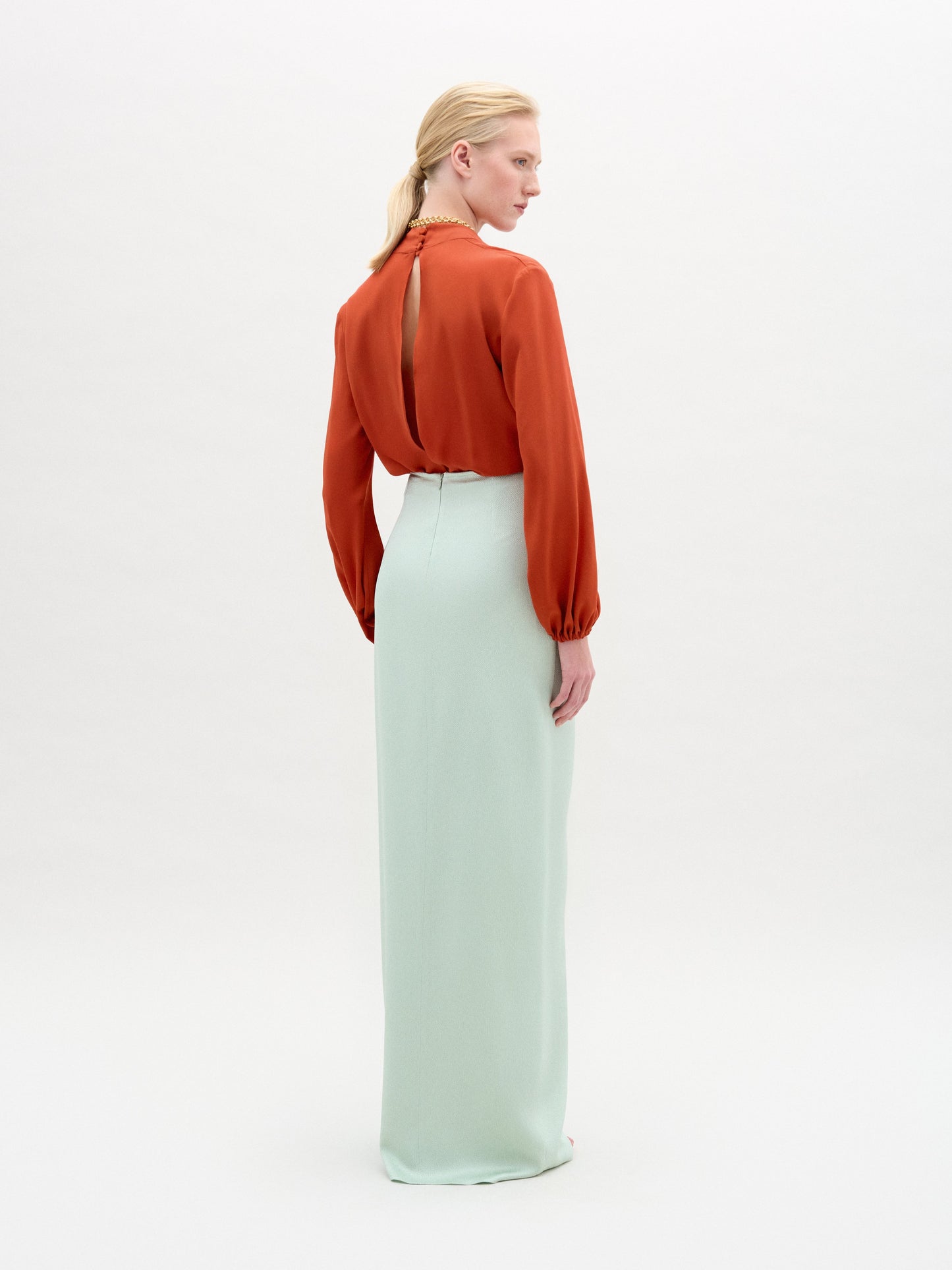 A person stands against a plain white background wearing the Gadea Skirt in light blue Italian fabric, an orange long-sleeve blouse, a gold belt, and a necklace.