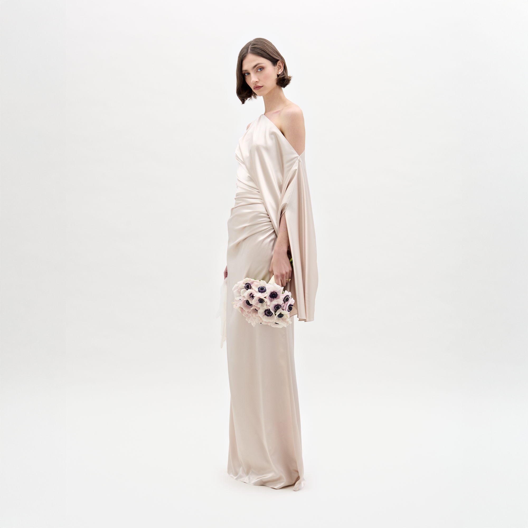 A woman in the SVT DESIGNS Dotty Dress, a long pale satin maxi with an asymmetric neckline and draped one-shoulder design, stands holding a bouquet of light-colored flowers against a plain white background.