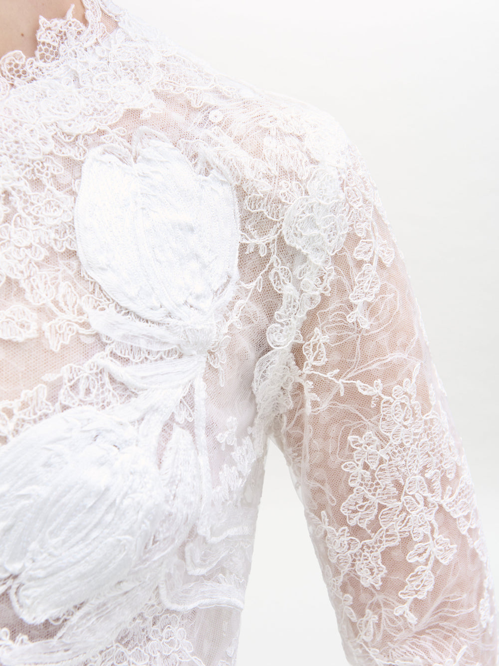Close-up of the Dava Dress by SVT DESIGNS, crafted from sheer white lace with floral patterns, intricate embroidery, and larger white floral appliqués. The delicate lace covers the shoulder and chest area.
