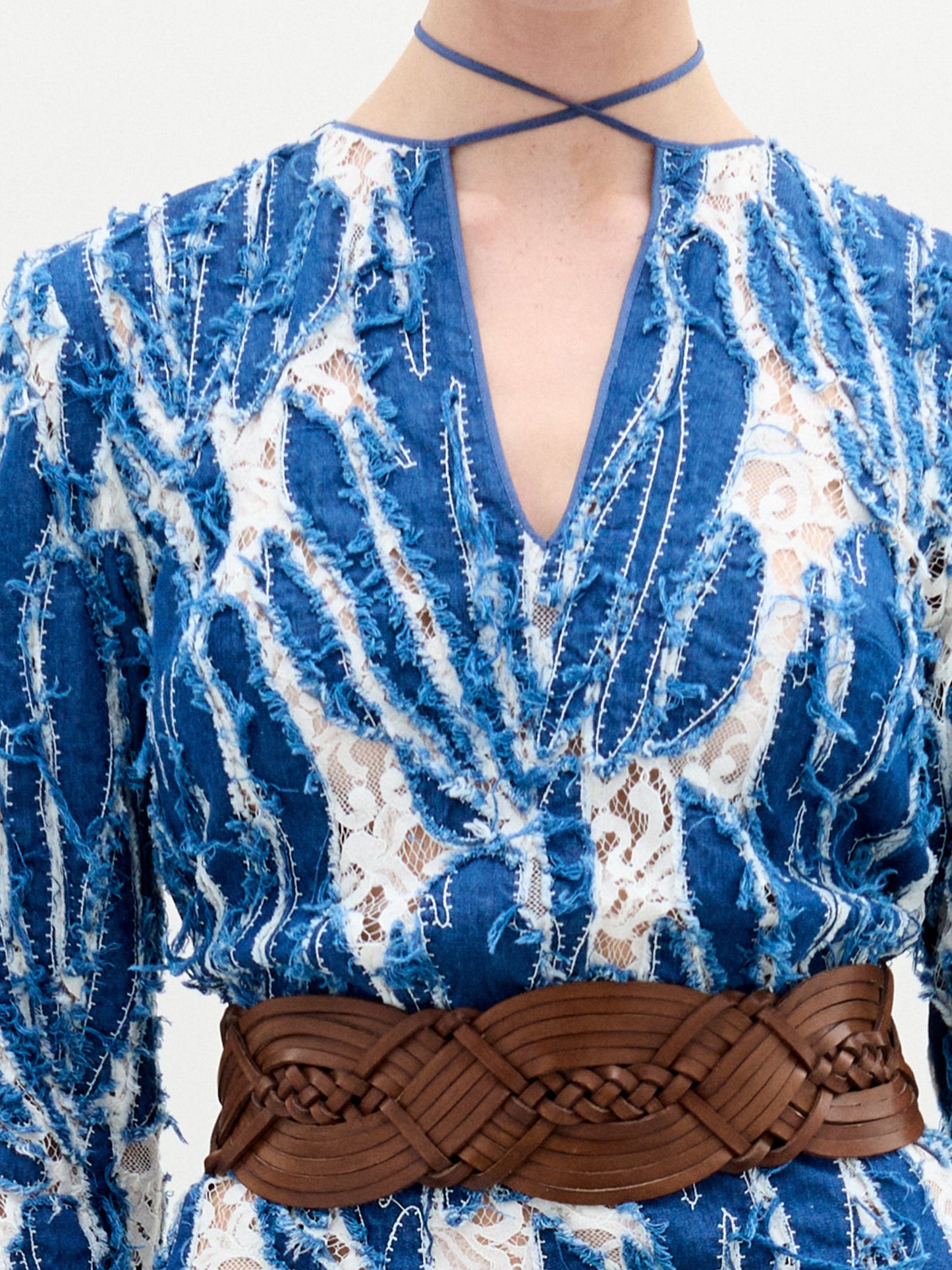 A person wears the SVT DESIGNS Ebba Dress, made of blue and white textured fabric with lace details, a V-neckline, leaf-like patterns, a dark brown braided belt, and a thin blue choker.