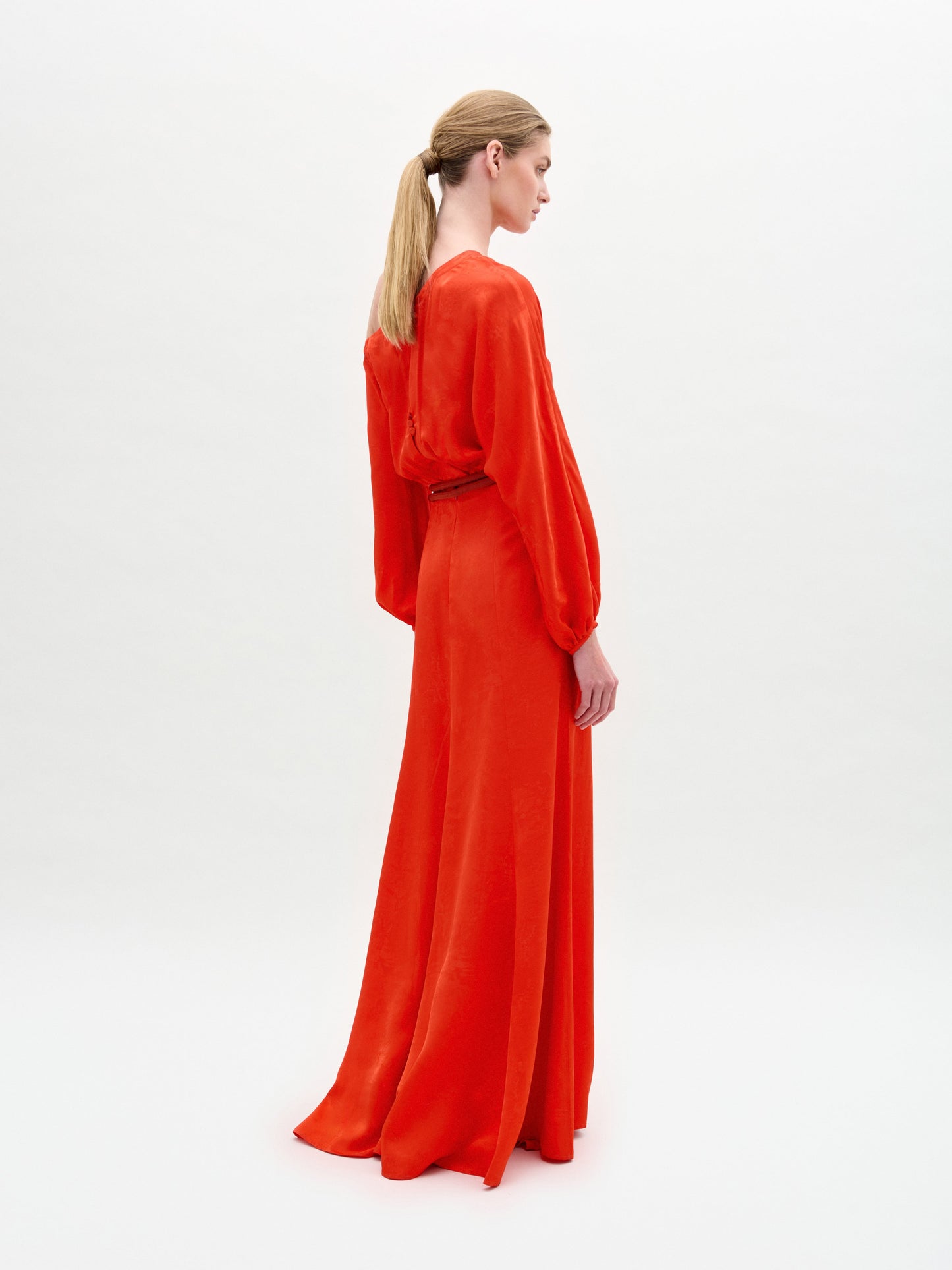 A woman with blond hair in a low ponytail models the Elga Dress, a long, bright orange maxi with long sleeves, crafted from fine Italian fabric. She stands against a plain white background, facing slightly away from the camera.