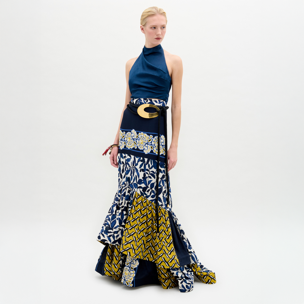 A person stands against a plain background wearing the Elliot Skirt by SVT DESIGNS, a patchwork maxi in blue, white, and yellow Italian fabric with geometric and floral patterns, paired with a blue halter top and wide gold-buckled belt.