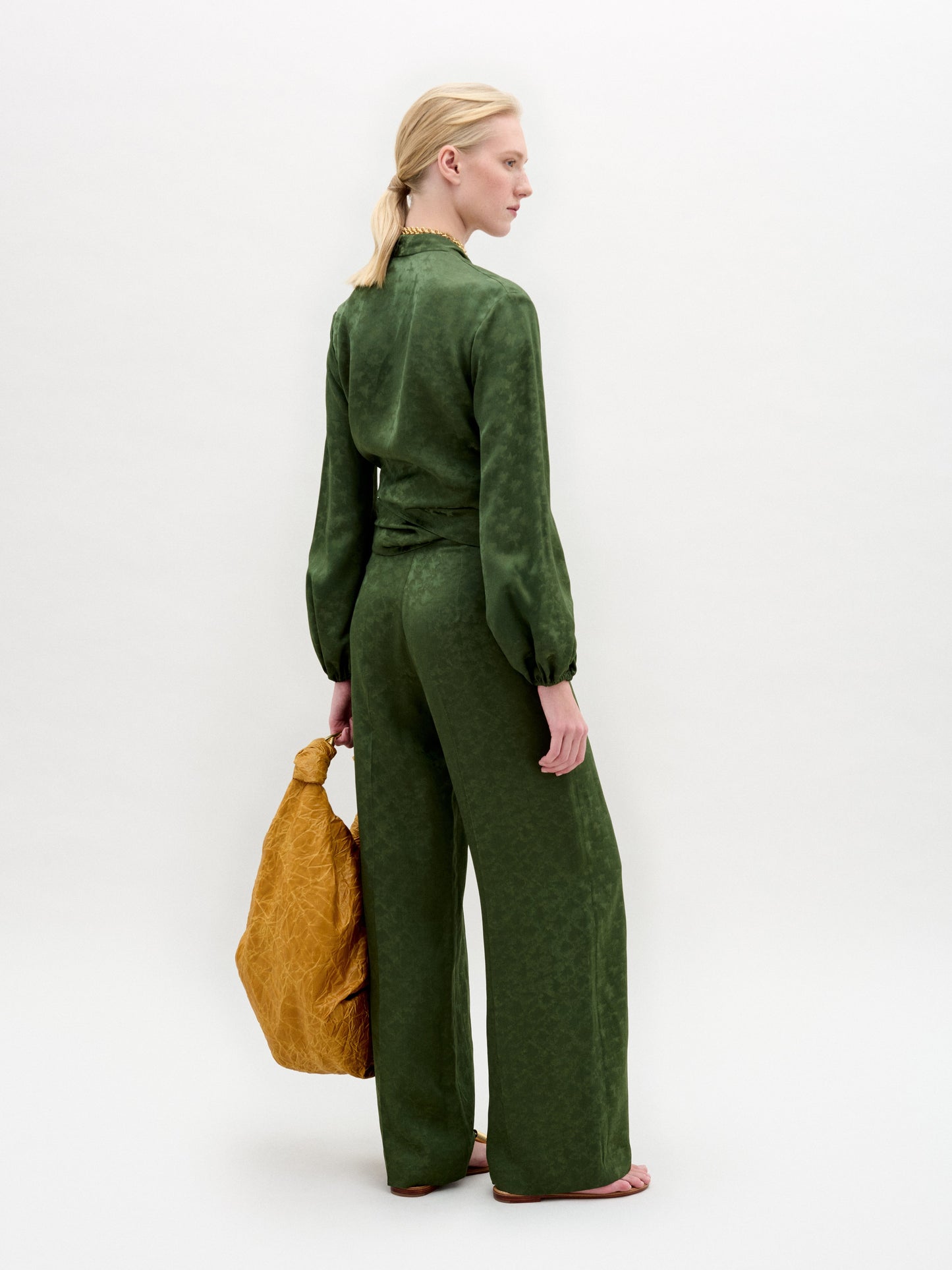 Against a plain backdrop, they wear the green Elvie Blouse by SVT DESIGNS with wrap-around ties and wide-leg pants, holding a large textured brown bag. Their hair is styled back as they gaze sideways with a neutral expression, highlighted by subtle gold embellishments.