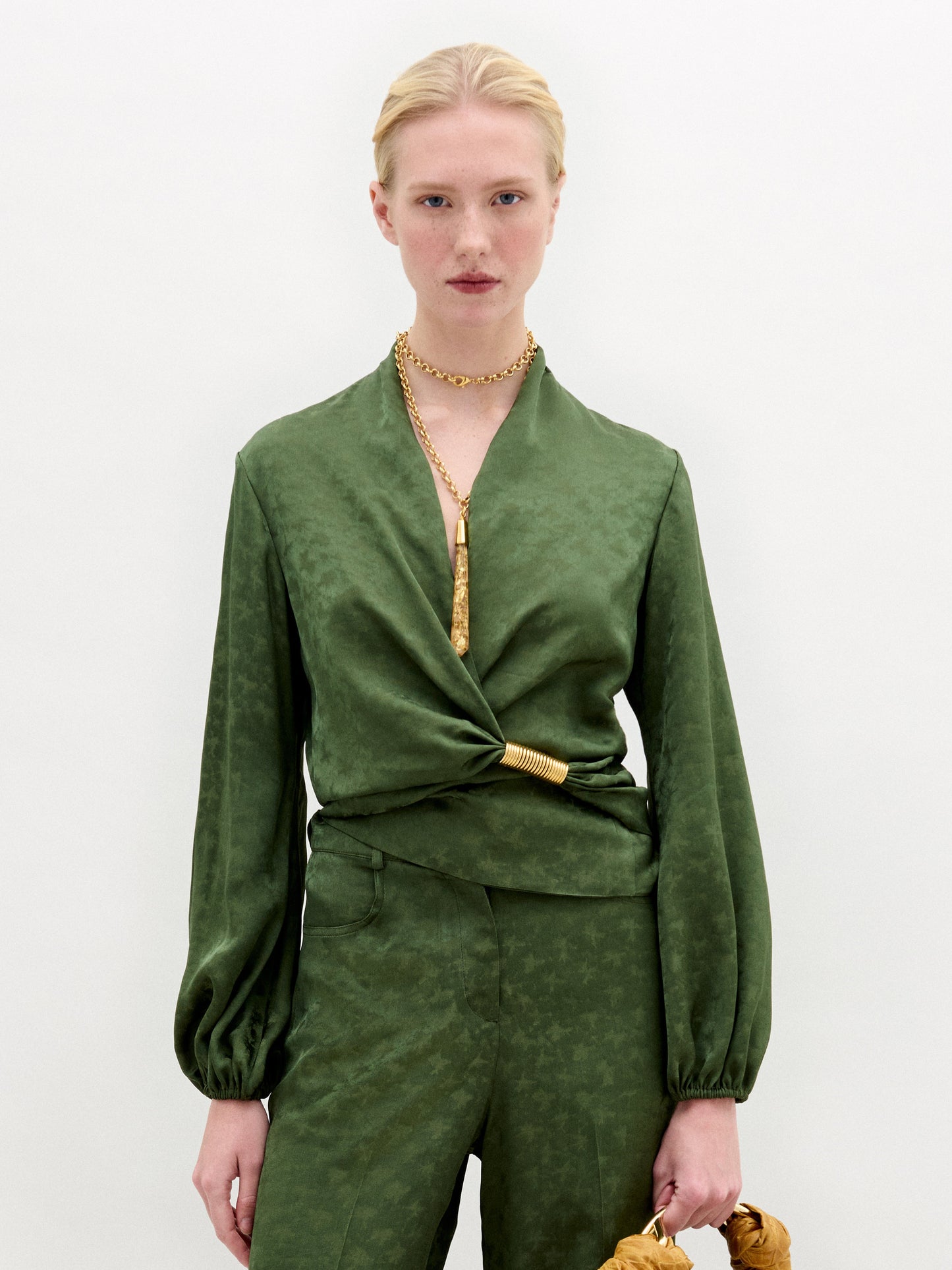 A person with light skin and blonde hair pulled back wears the SVT DESIGNS Elvie Blouse, a green long-sleeved wrap top with matching pants, standing against a white background, accessorized with gold jewelry and a gold waist detail.