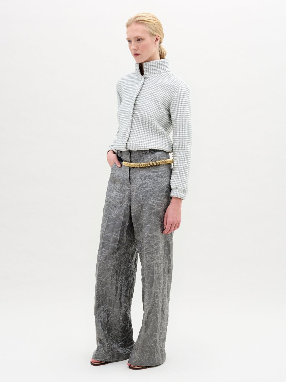 A person stands against a plain background wearing a textured light gray zip-up jacket and SVT DESIGNS Emine Pant, high-waisted wide-leg crinkled gray pants with hands in pockets, proudly made in Colombia.