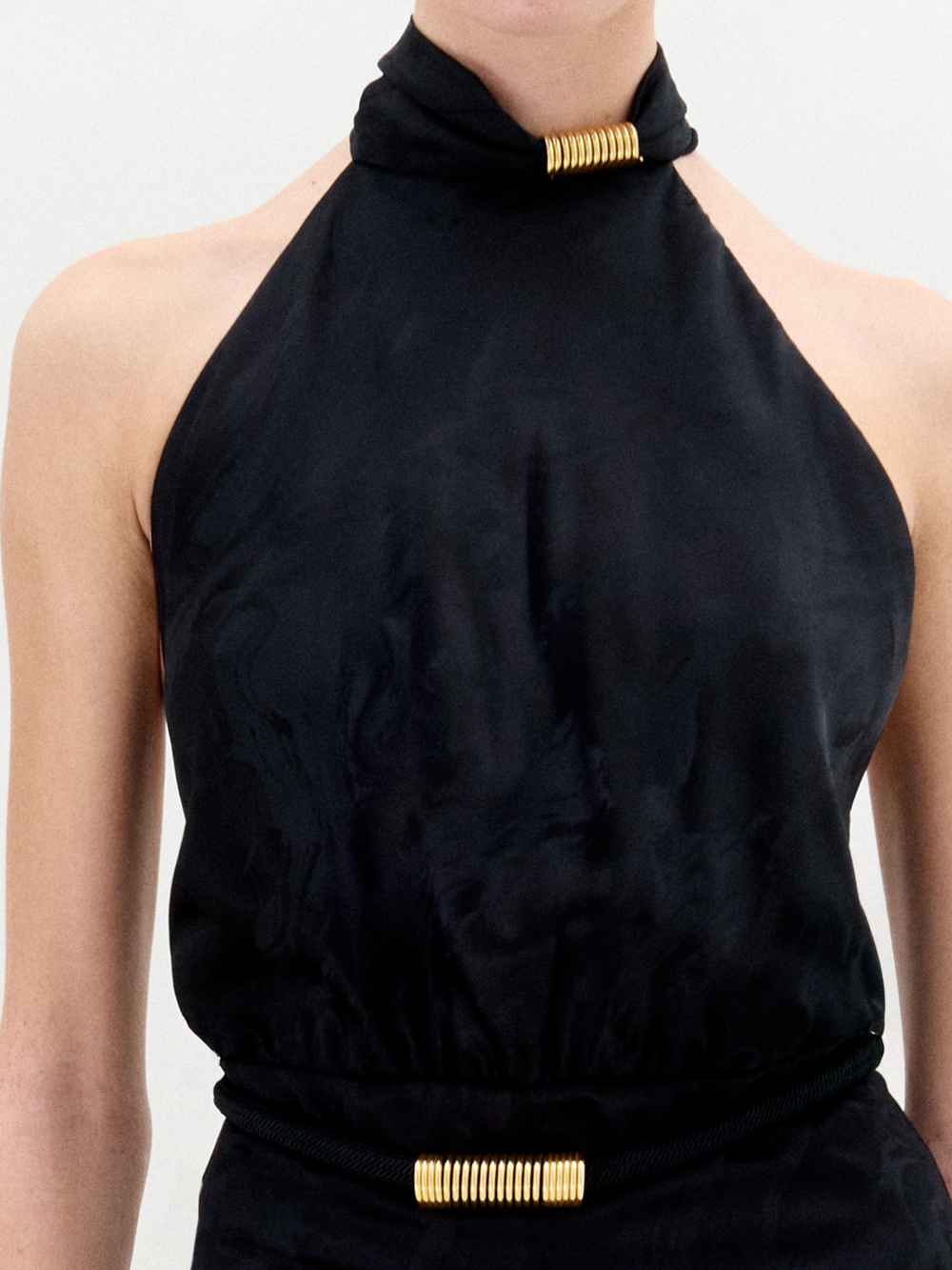 A person wears the Enara Jumpsuit by SVT DESIGNS—a sleeveless black halter-neck piece with gold cylindrical accents at the neck and waist, crafted from subtly sheened Italian fabric with gathered detail. The background is plain and light-colored.