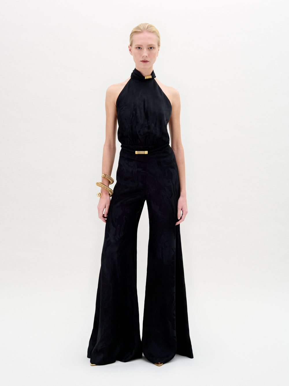 A person stands against a white background wearing the Enara Jumpsuit by SVT DESIGNS, a sleeveless black halter-neck style with wide legs made from Italian fabric, paired with gold bracelets and black pointed shoes. Their expression is neutral, arms relaxed.