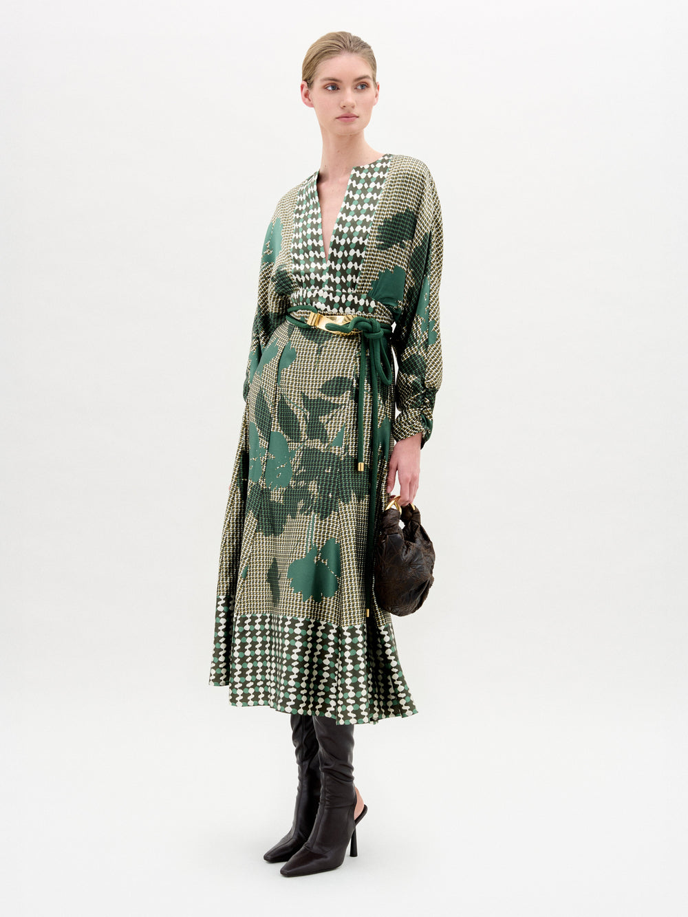 A woman stands against a plain background wearing the SVT DESIGNS Erin Skirt in green Italian fabric, paired with black knee-high boots and a small dark handbag. Her hair is pulled back.