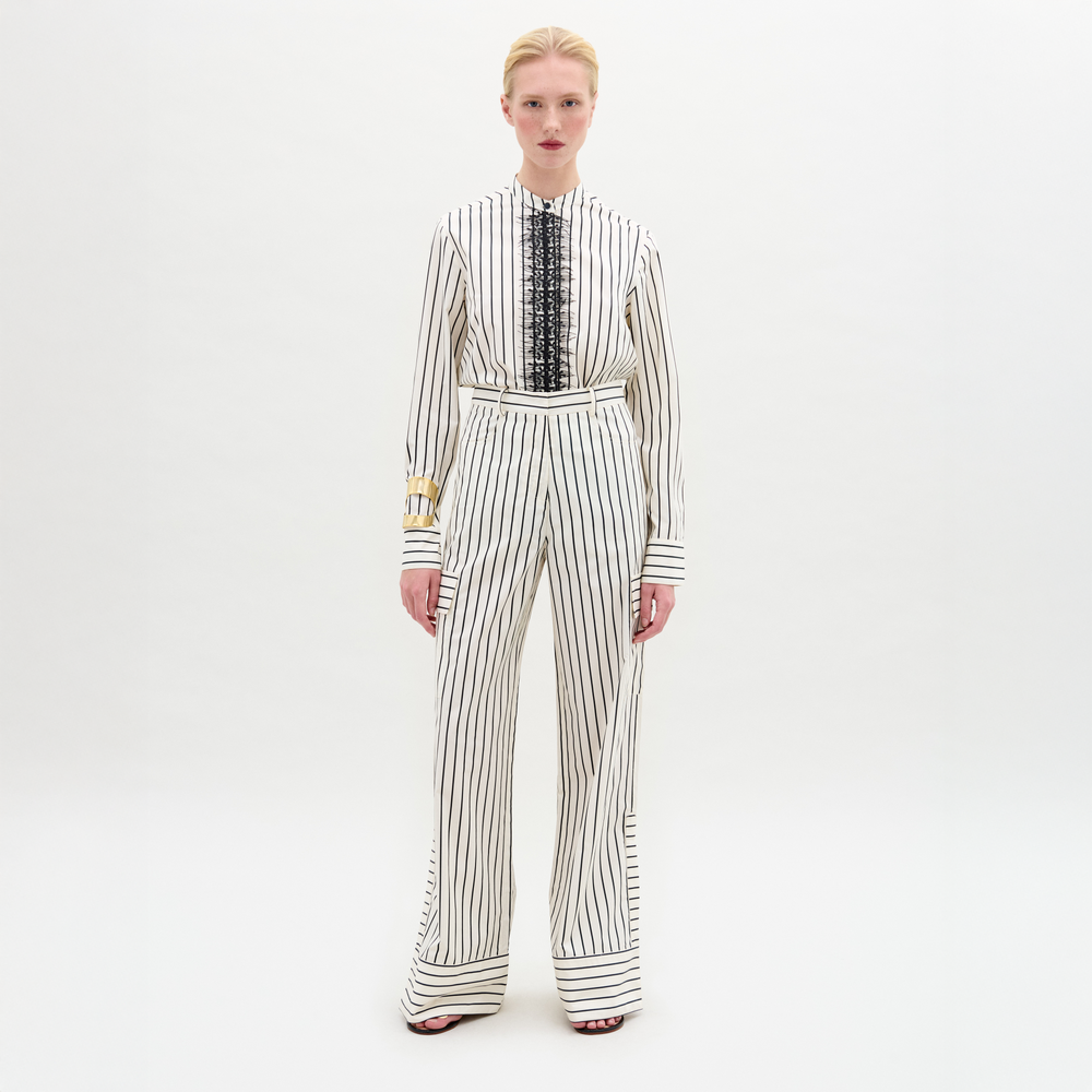 A person stands against a plain white background wearing the SVT DESIGNS Falak Pant, a wide-leg, long-sleeved white jumpsuit with vertical black stripes and front black lace detailing, crafted from premium Italian fabric.