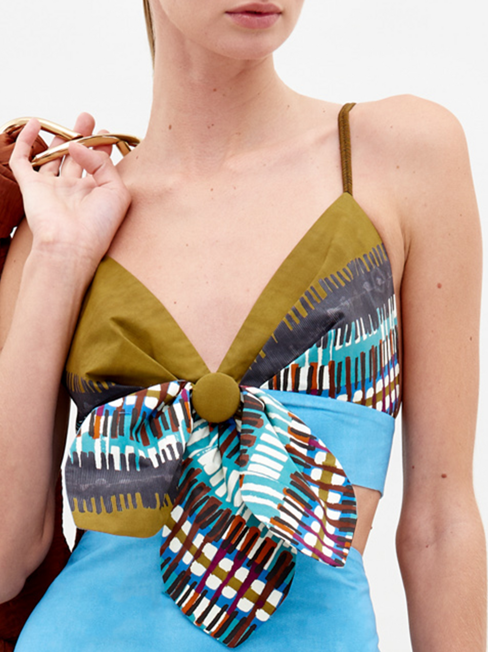 A person wearing the Flor Top by SVT DESIGNS—a sleeveless piece with olive green straps, a deep V neckline, a large front bow detail, and blue, brown, and white Italian-inspired geometric patterns.