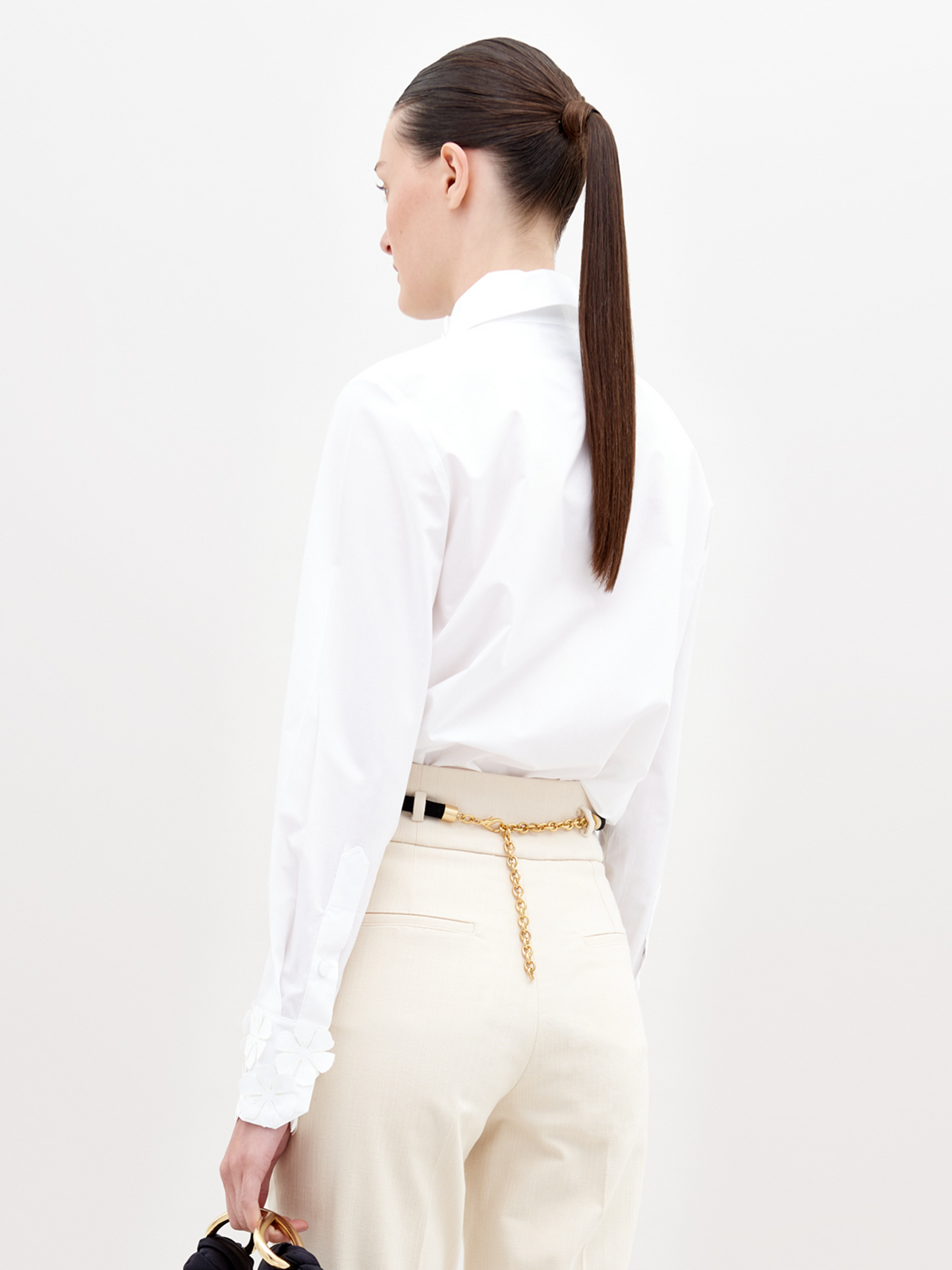 A woman with straight brown hair in a low ponytail models the Flower Blouse, made from 100% cotton, paired with beige pants and a gold chain belt. She stands against a plain light background, shown from the back and slightly to the side.