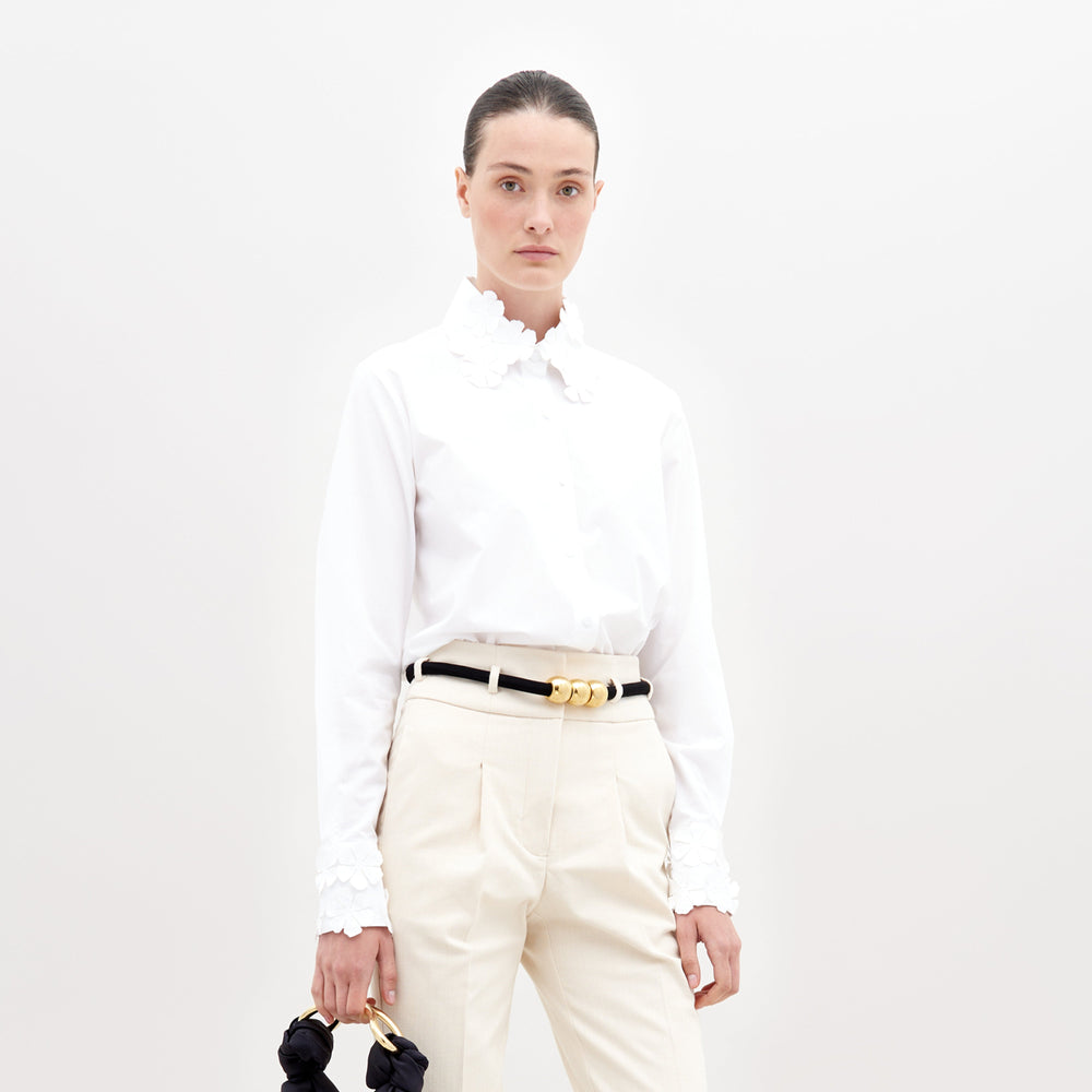 A person stands against a plain white background wearing the SVT DESIGNS Flower Blouse (100% cotton, floral appliqué cuffs), cream trousers, a black and gold belt, and holding a small black item in their left hand.
