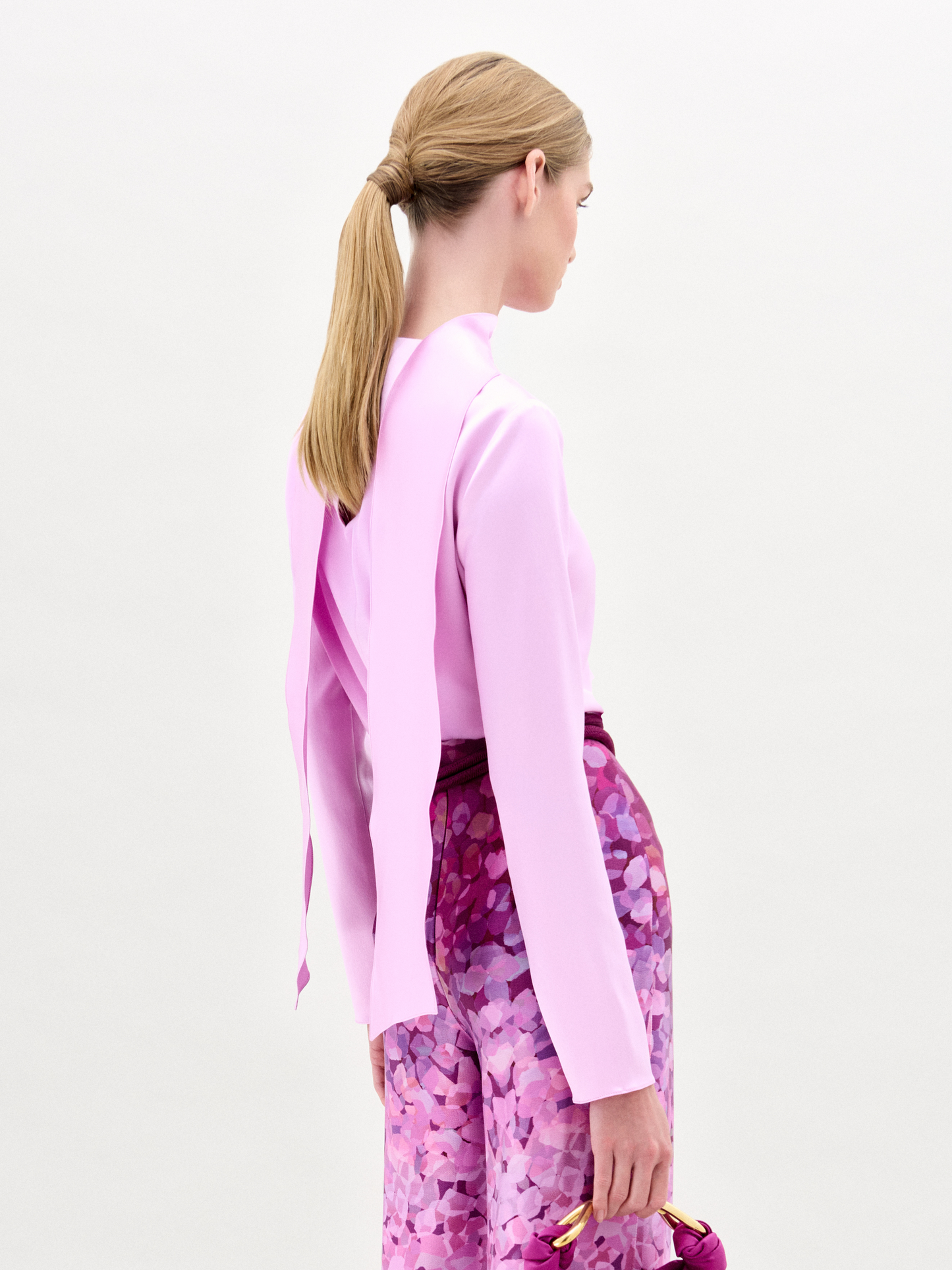 A woman poses against a plain background in the Gaby Blouse, a light pink silk long-sleeve top with a front slit, paired with high-waisted pink and purple patterned pants, a wide belt, and a small matching handbag.
