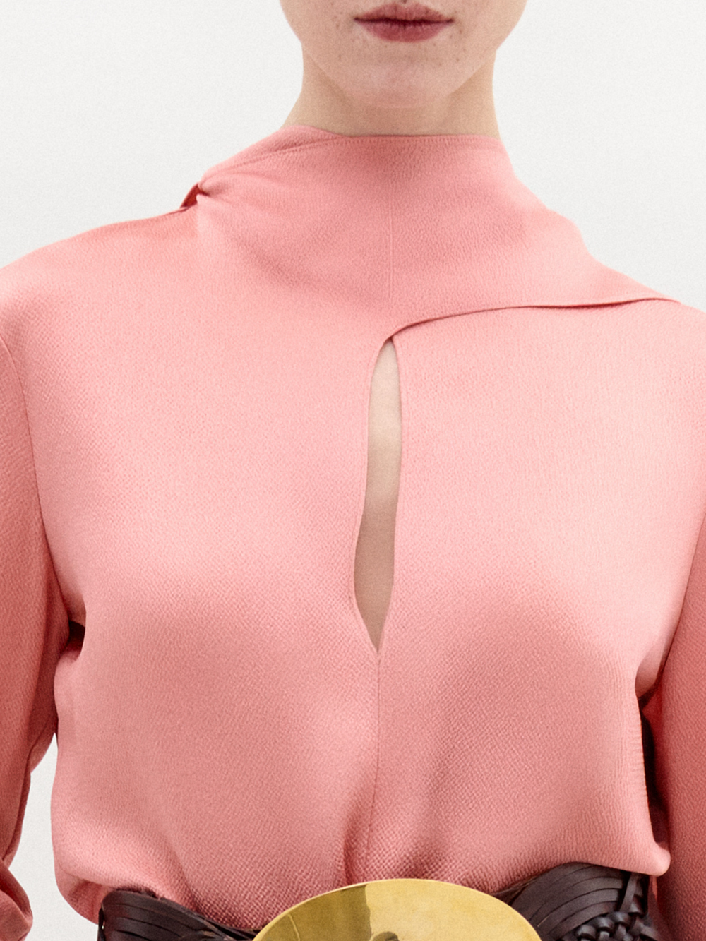 A person wears the SVT DESIGNS Gaby Blouse—a textured pink high-neck top with a vertical front slit. The cropped image shows part of a brown belt with a large gold buckle.