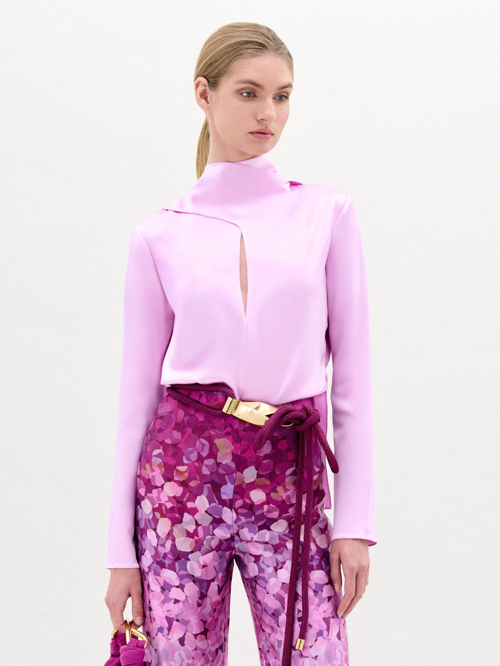 A woman poses against a plain light background in the SVT DESIGNS Gaby Blouse—a high-neck silk top with a front slit—paired with high-waisted purple patterned pants and a small pink handbag. This chic look is made in Colombia.
