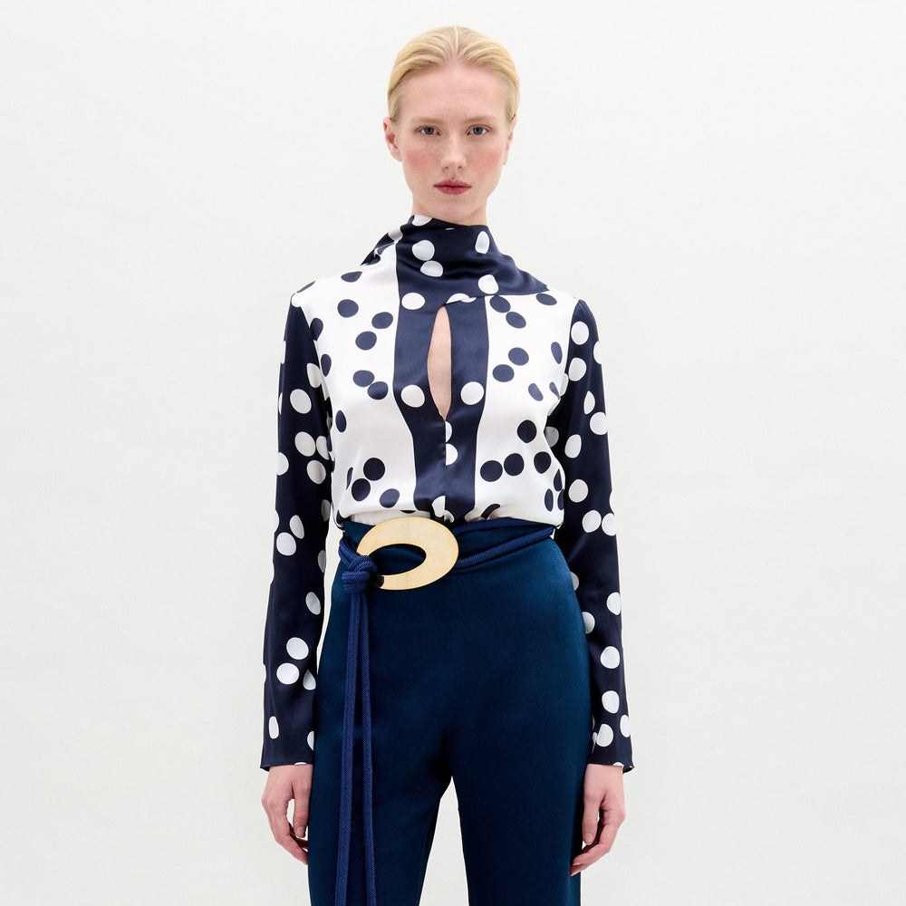 A blonde model wears the SVT DESIGNS Gaby Blouse—a navy and white polka dot silk top with a high neck, long sleeves, and keyhole cutout—paired with dark blue pants and a wide belt with a large round buckle. Plain light background.