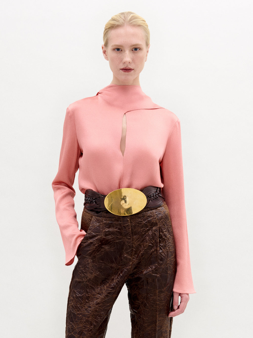 A person stands against a plain white background wearing the SVT DESIGNS Gaby Blouse in pale pink, featuring a high neck and keyhole cutout, styled with brown Italian fabric pants and a wide belt with a shiny gold buckle.