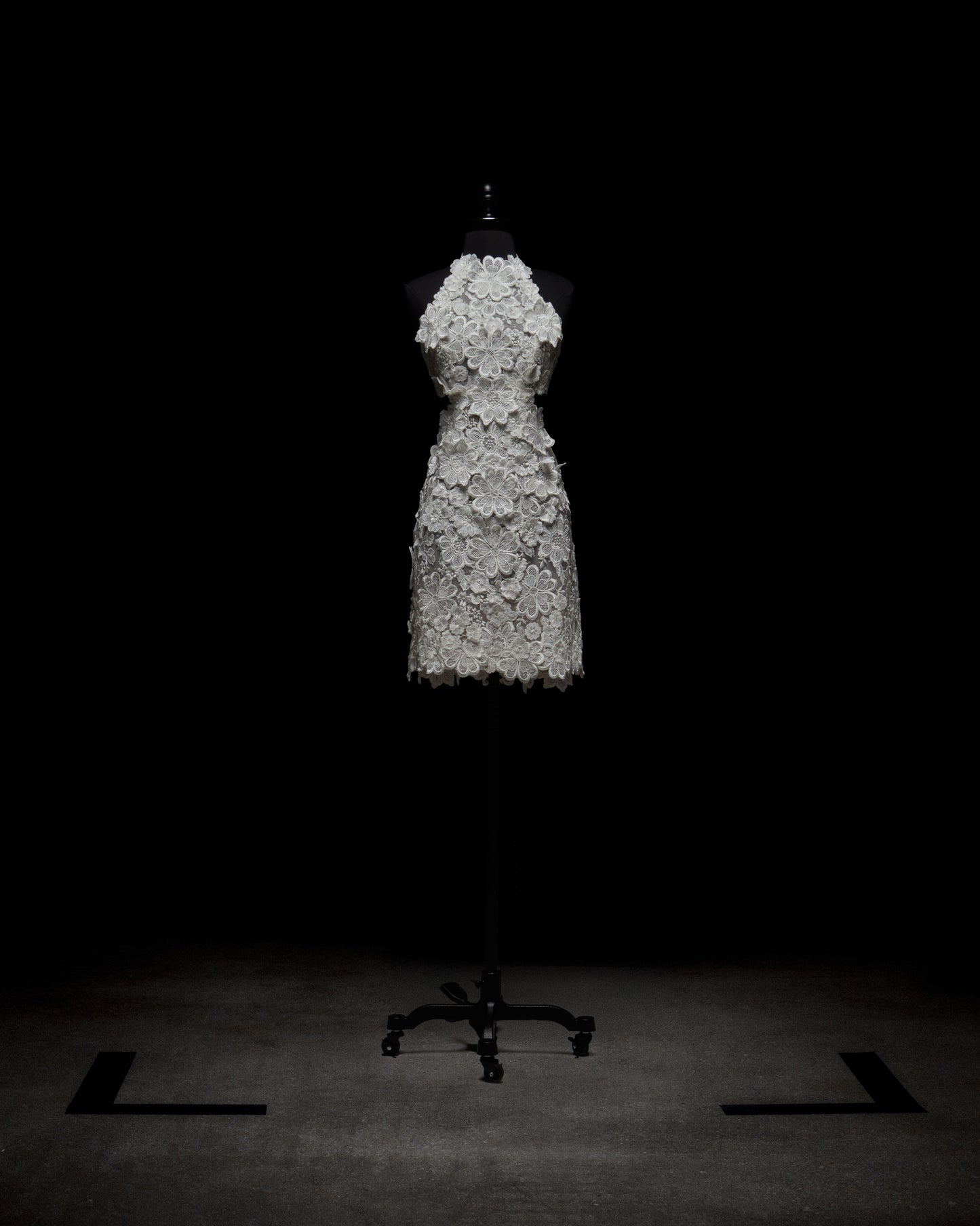 A mannequin showcases the Garazi Dress by SVT DESIGNS, a sleeveless, knee-length white dress with intricate lace floral patterns against a dark backdrop. It's displayed on a mobile black stand at the image's center.