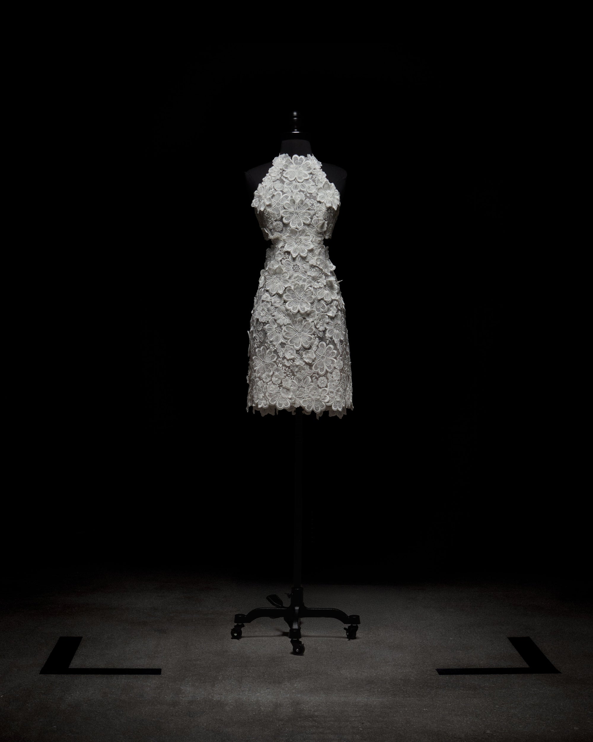 A mannequin showcases the Garazi Dress by SVT DESIGNS, a sleeveless, knee-length white dress with intricate lace floral patterns against a dark backdrop. It's displayed on a mobile black stand at the image's center.