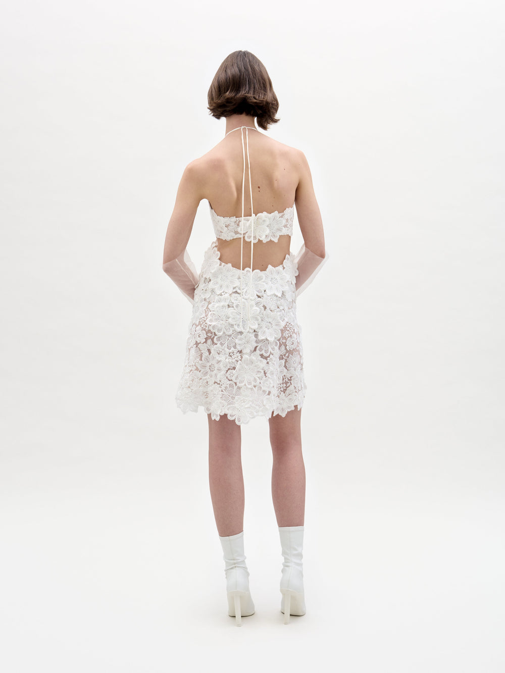 Against a plain background, an individual models the Garazi Dress by SVT DESIGNS, featuring a white floral lace halter design with an open back, paired with white heeled boots and sheer gloves.