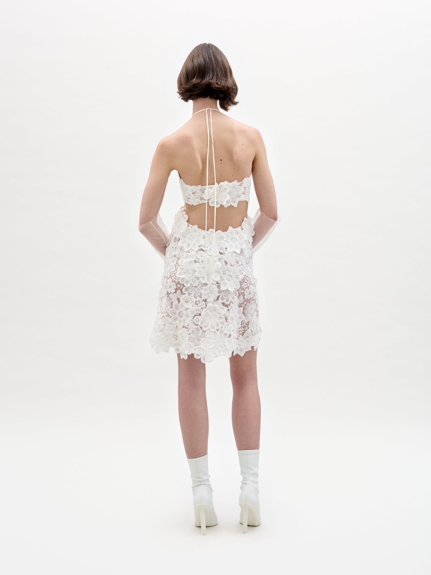 Against a plain background, an individual models the Garazi Dress by SVT DESIGNS, featuring a white floral lace halter design with an open back, paired with white heeled boots and sheer gloves.