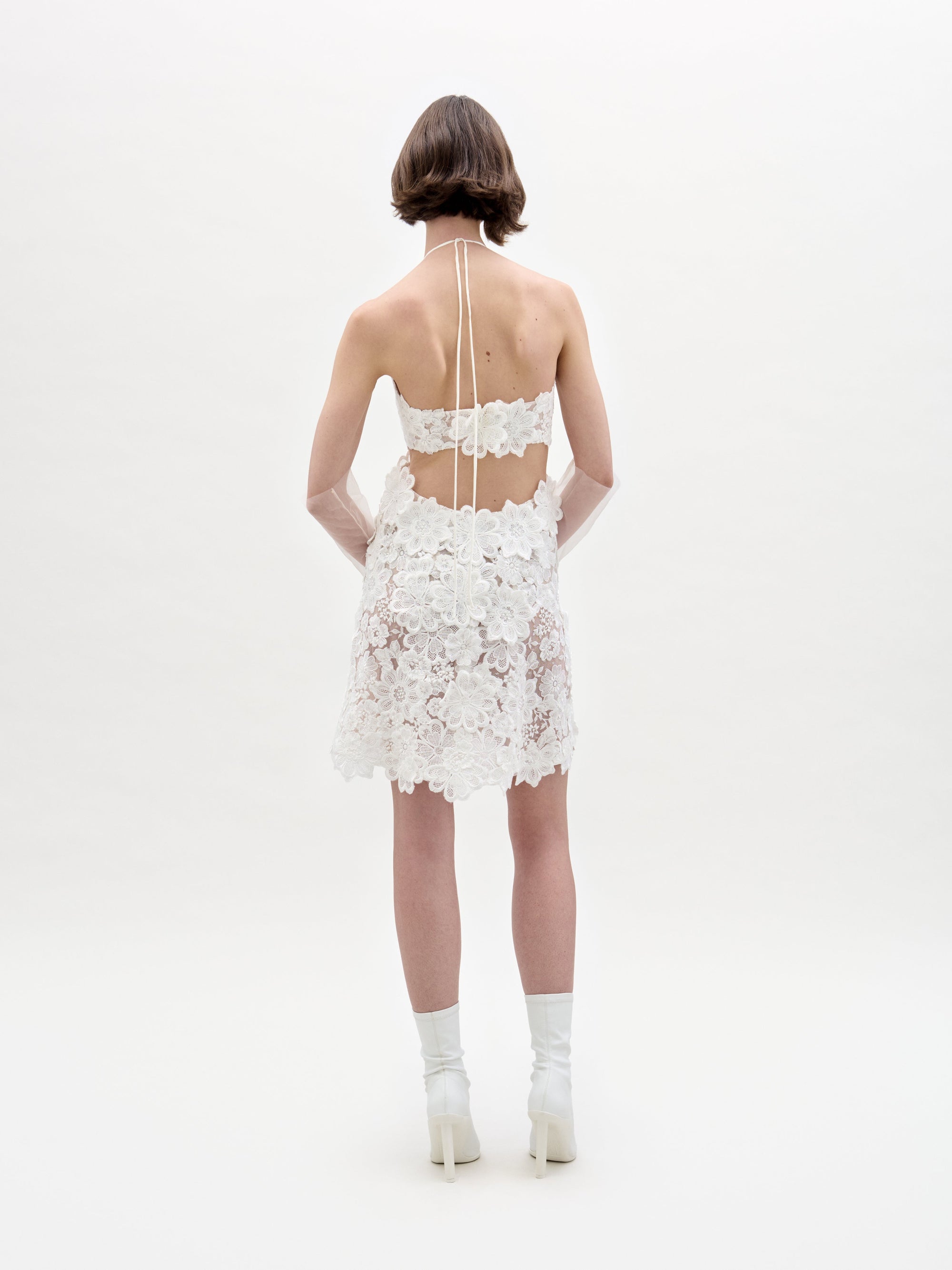 Against a plain background, an individual models the Garazi Dress by SVT DESIGNS, featuring a white floral lace halter design with an open back, paired with white heeled boots and sheer gloves.