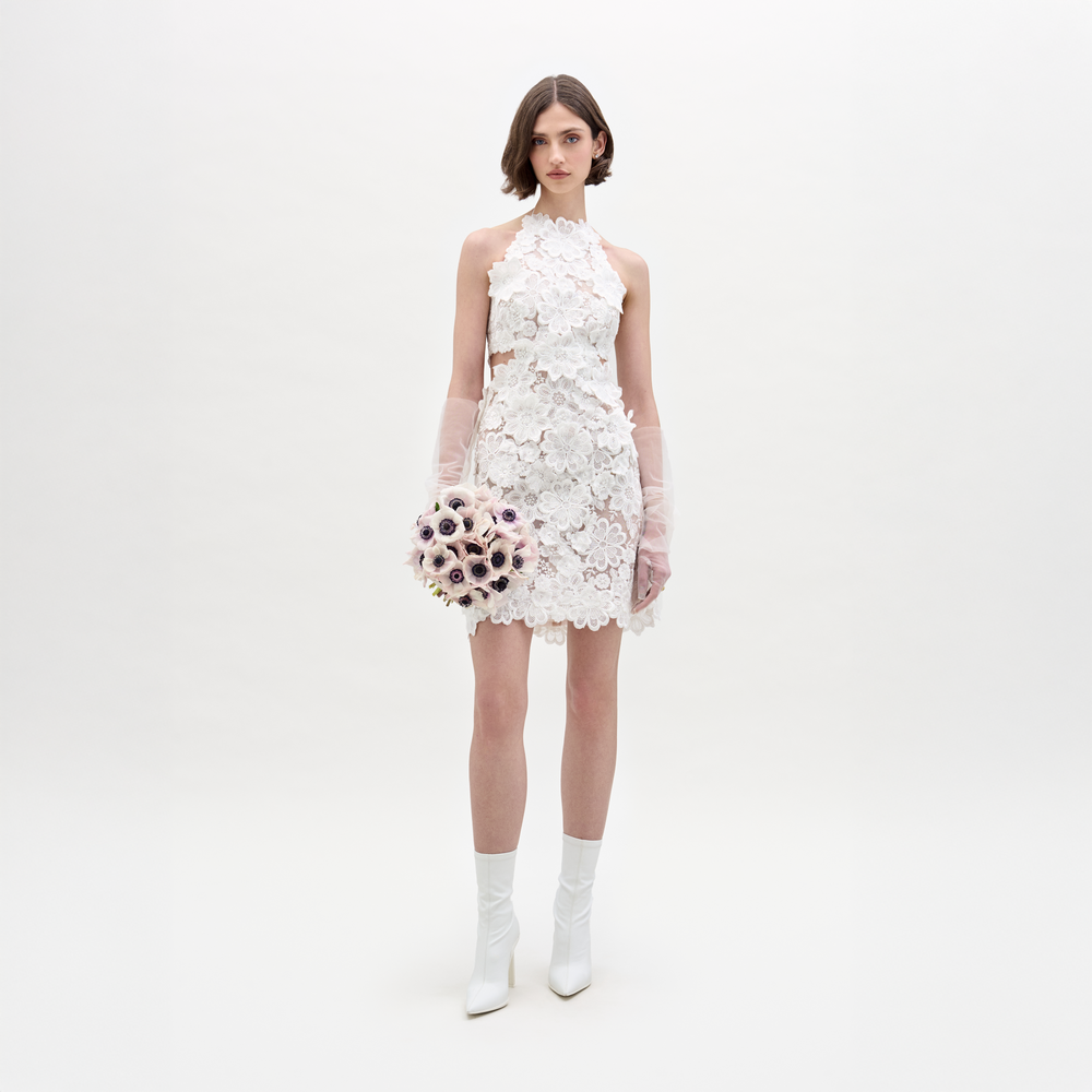 A woman wears the SVT DESIGNS Garazi Dress, a sleeveless white lace dress with floral appliqué, paired with matching boots and translucent gloves. She holds a bouquet and faces the camera against a plain white background.