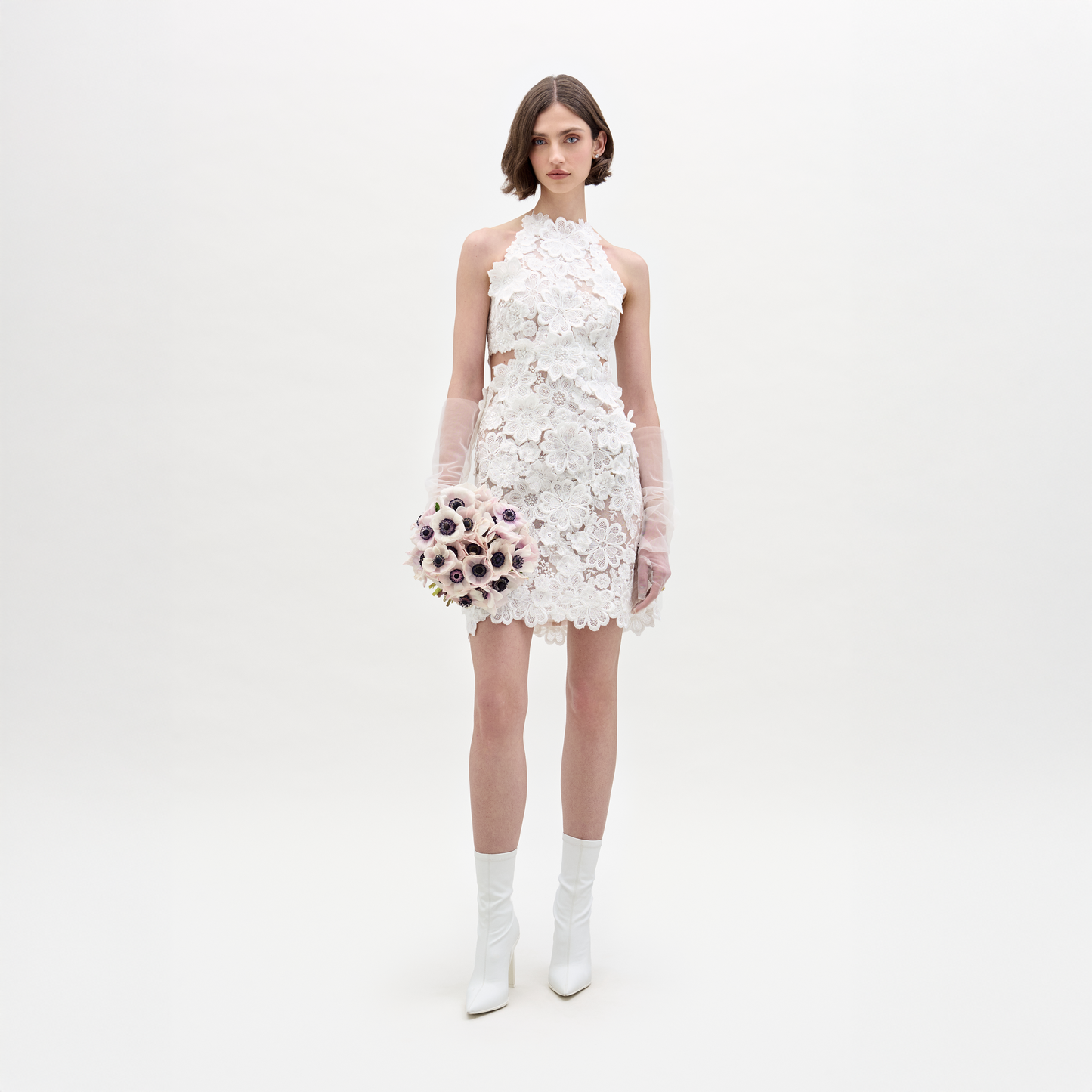 A woman wears the SVT DESIGNS Garazi Dress, a sleeveless white lace dress with floral appliqué, paired with matching boots and translucent gloves. She holds a bouquet and faces the camera against a plain white background.