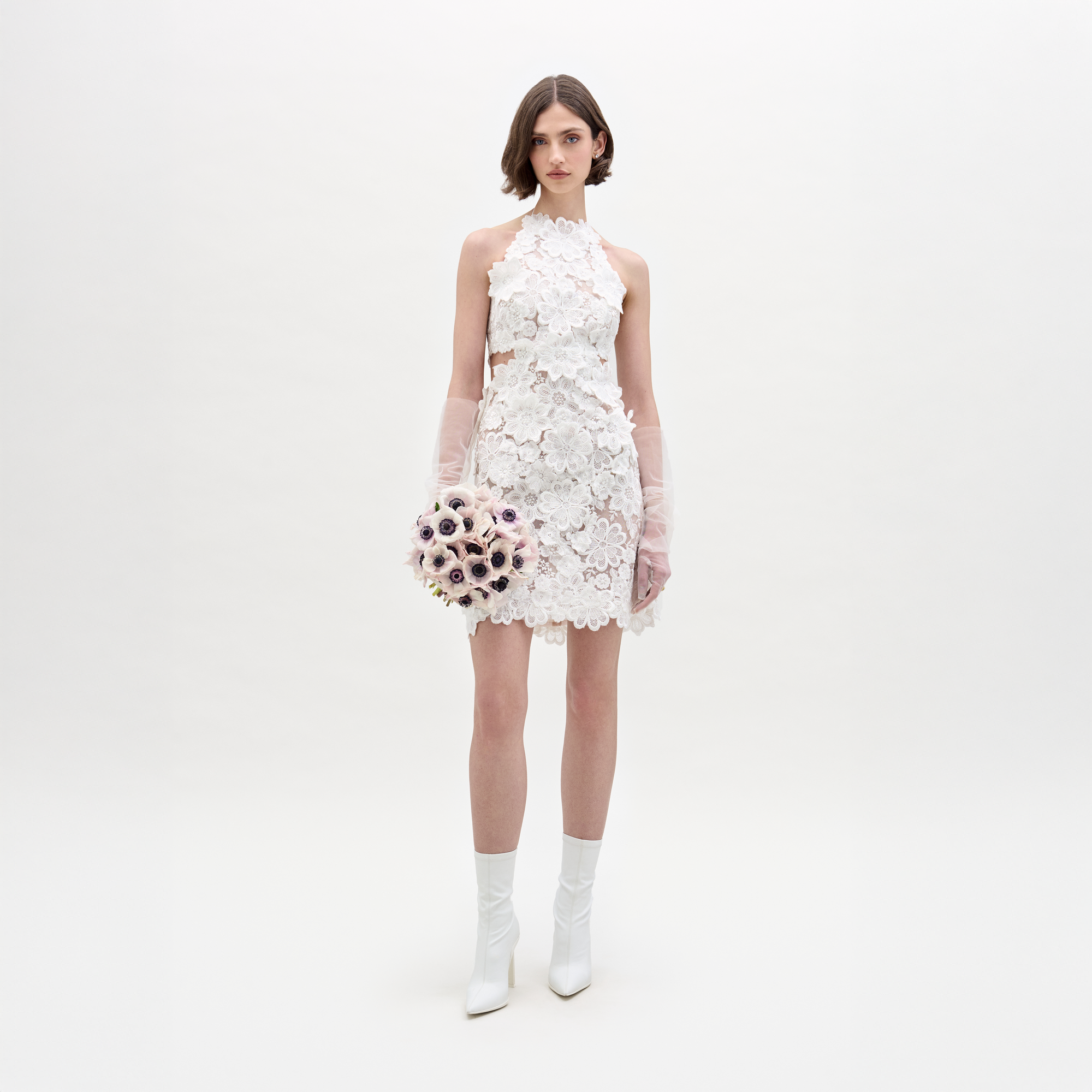 A woman wears the SVT DESIGNS Garazi Dress, a sleeveless white lace dress with floral appliqué, paired with matching boots and translucent gloves. She holds a bouquet and faces the camera against a plain white background.