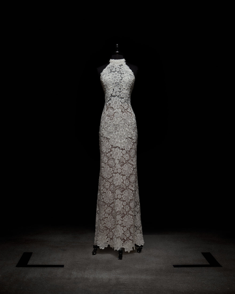 The Gardenia Dress by SVT DESIGNS, a long sleeveless creation of detailed white lace with a high neckline and floral patterns, is showcased on a mannequin against a dark backdrop.
