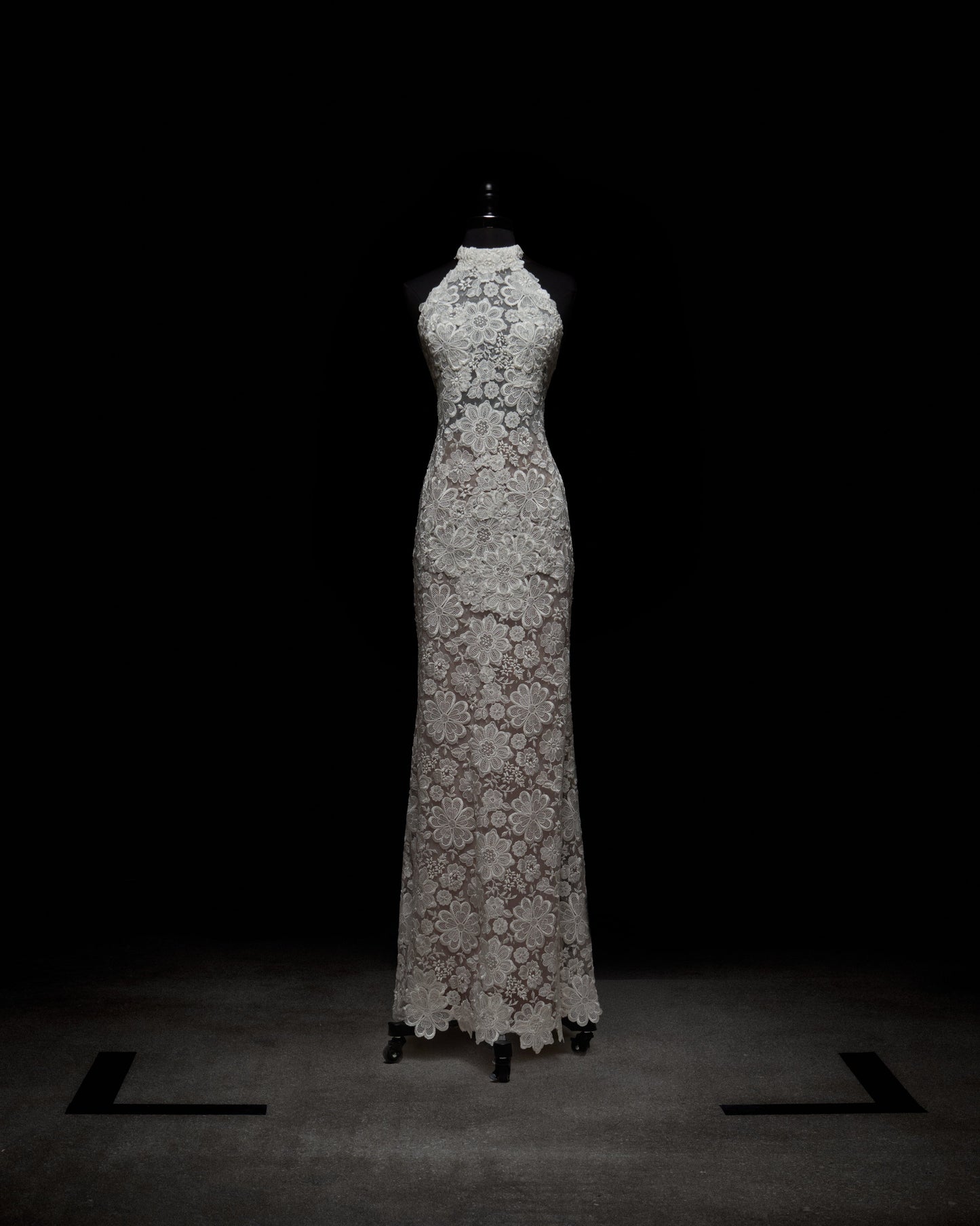 The Gardenia Dress by SVT DESIGNS, a long sleeveless creation of detailed white lace with a high neckline and floral patterns, is showcased on a mannequin against a dark backdrop.