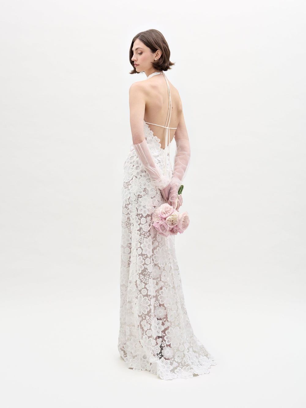 A woman in SVT DESIGNS' Gardenia Dress, a lace bridal gown with a halter-neck and open back, stands against a white background. She holds a bouquet of pink roses behind her with both hands while wearing sheer gloves. Her hair is short and styled simply.
