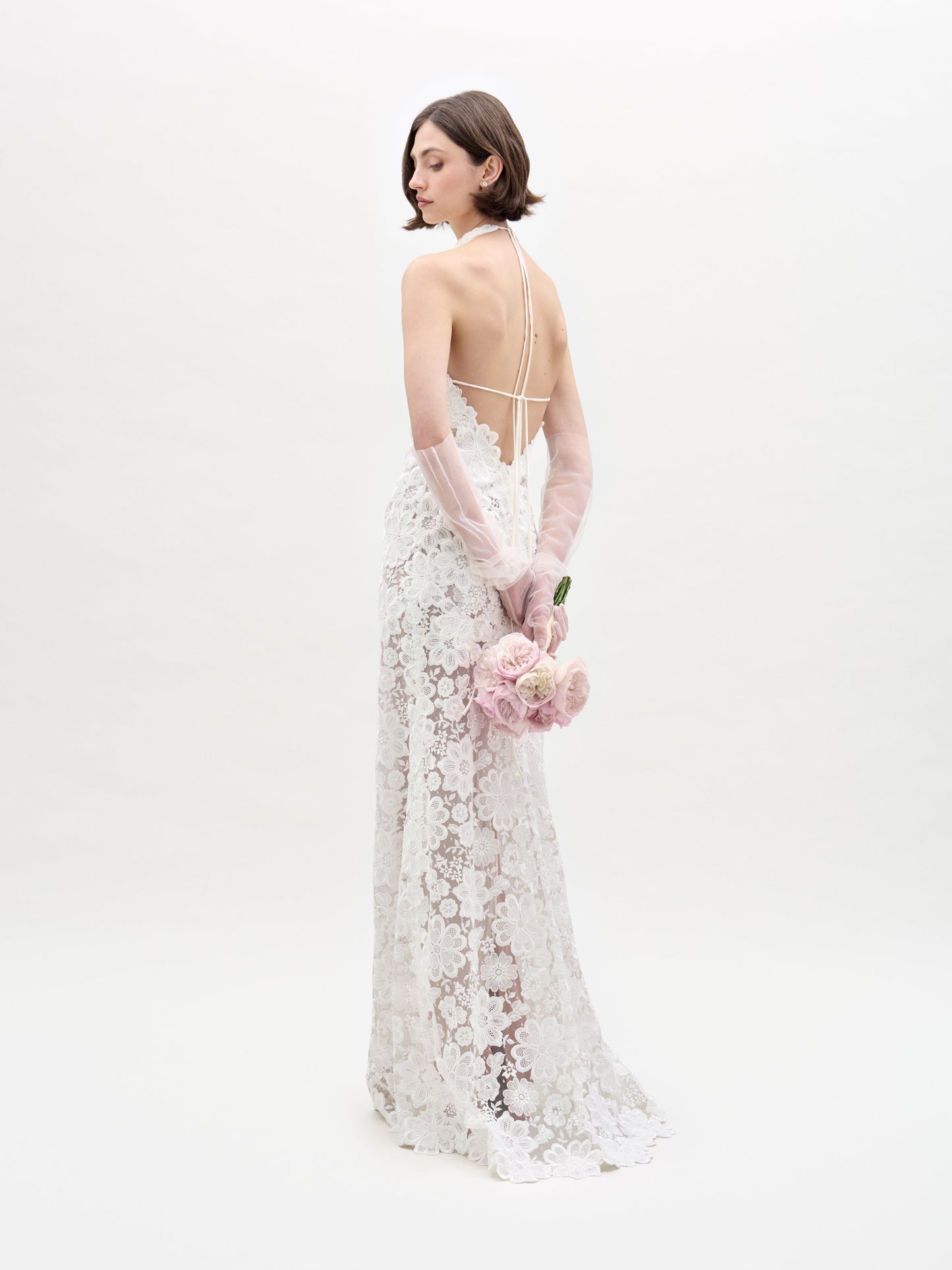 A woman in SVT DESIGNS' Gardenia Dress, a lace bridal gown with a halter-neck and open back, stands against a white background. She holds a bouquet of pink roses behind her with both hands while wearing sheer gloves. Her hair is short and styled simply.