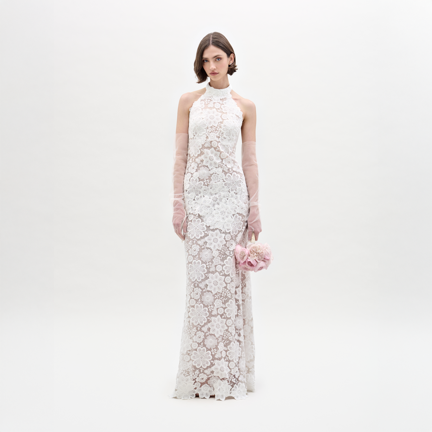 A woman poses against a plain white backdrop in the SVT DESIGNS Gardenia Dress, a sleeveless high-neck white sheer lace maxi with floral patterns and open back. She holds a pink bouquet and wears long, sheer gloves.