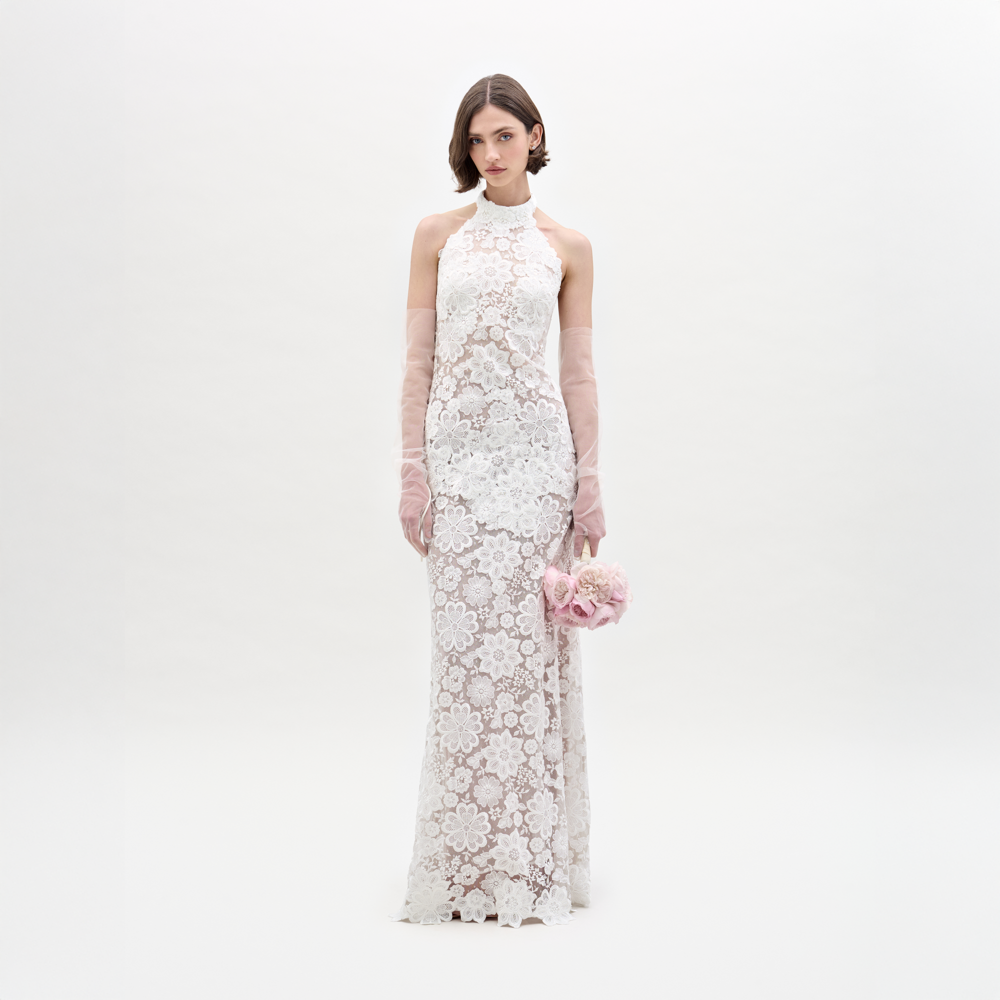 A woman poses against a plain white backdrop in the SVT DESIGNS Gardenia Dress, a sleeveless high-neck white sheer lace maxi with floral patterns and open back. She holds a pink bouquet and wears long, sheer gloves.