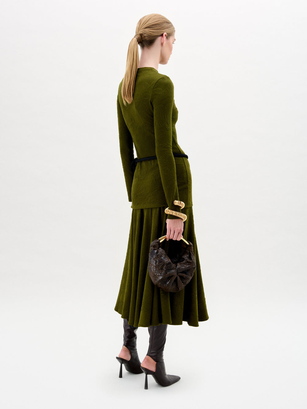 A woman with blonde hair in a low ponytail wears SVT DESIGNS’ olive green Gilda Skirt and matching long-sleeve top, styled with a black belt, brown heeled boots, and a textured dark brown handbag against a plain background.