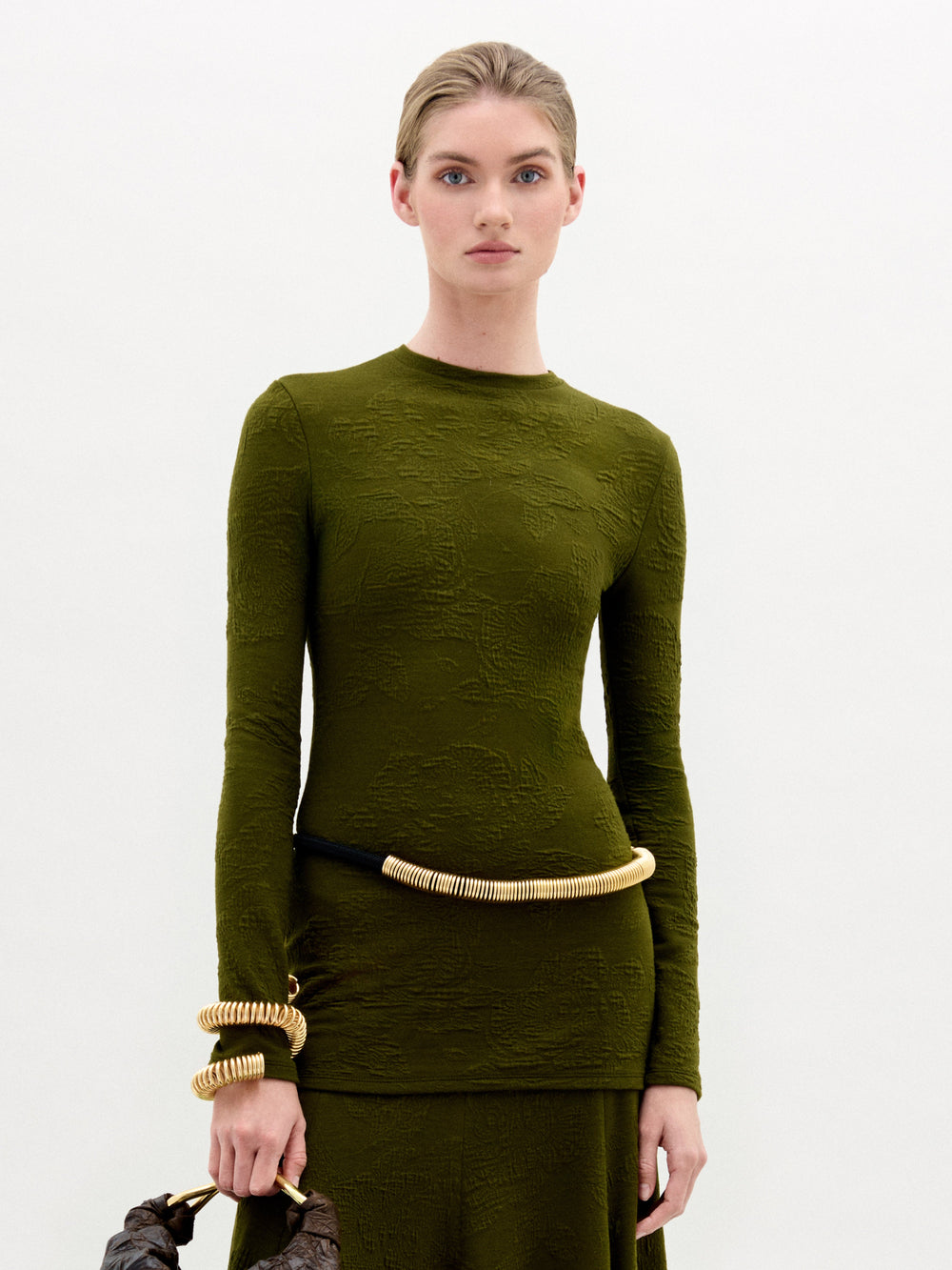 A person with light skin and blonde hair pulled back wears the Gen Tshirt by SVT DESIGNS. They accessorize with gold coil jewelry on their wrist and waist, holding a wooden-handle bag. The background is plain white.