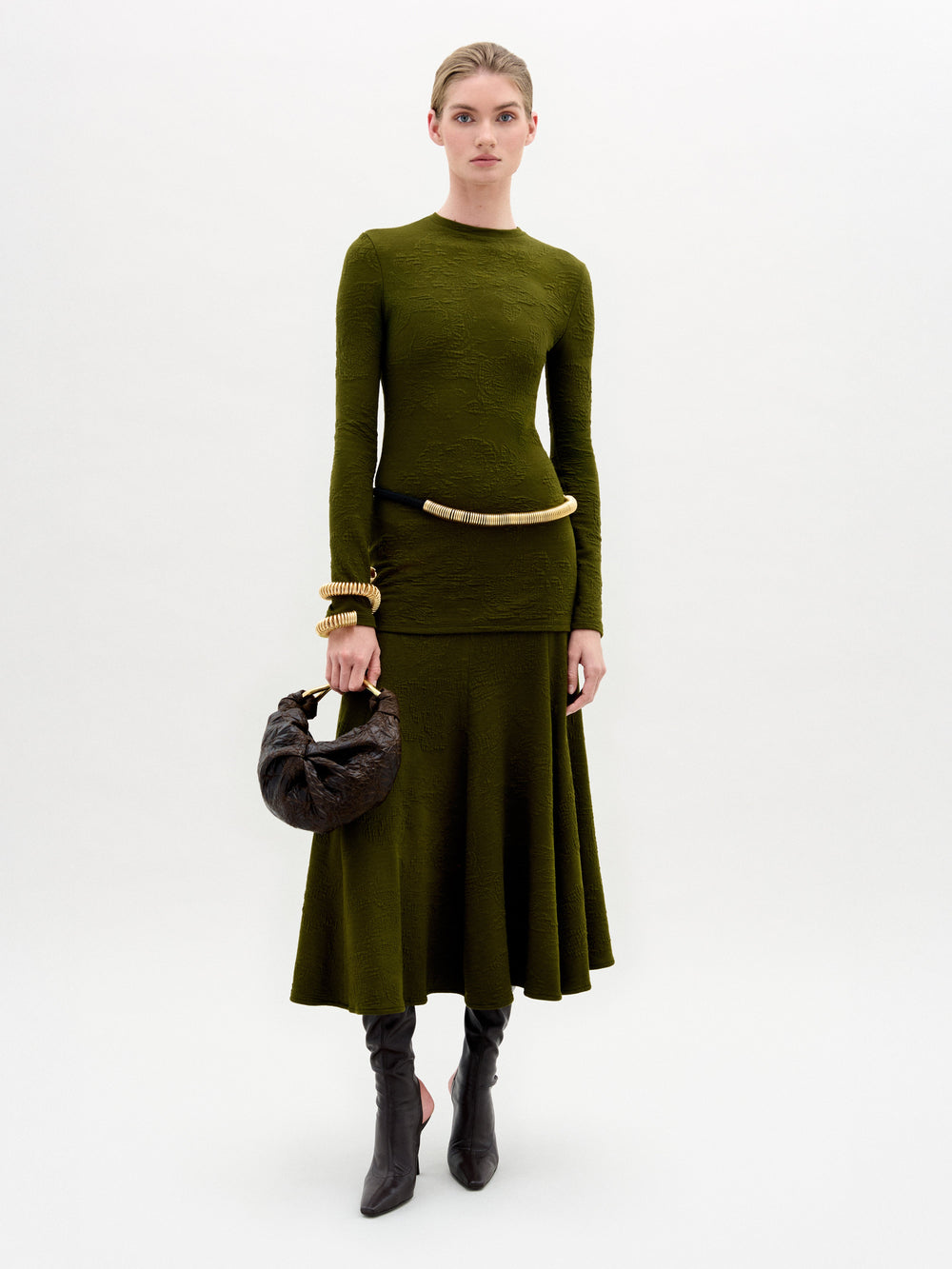 A woman stands against a white background wearing the SVT DESIGNS Gilda Skirt in olive green, styled with long sleeves, black knee-high boots, gold bangles, a curved belt, and holding a textured dark brown handbag.
