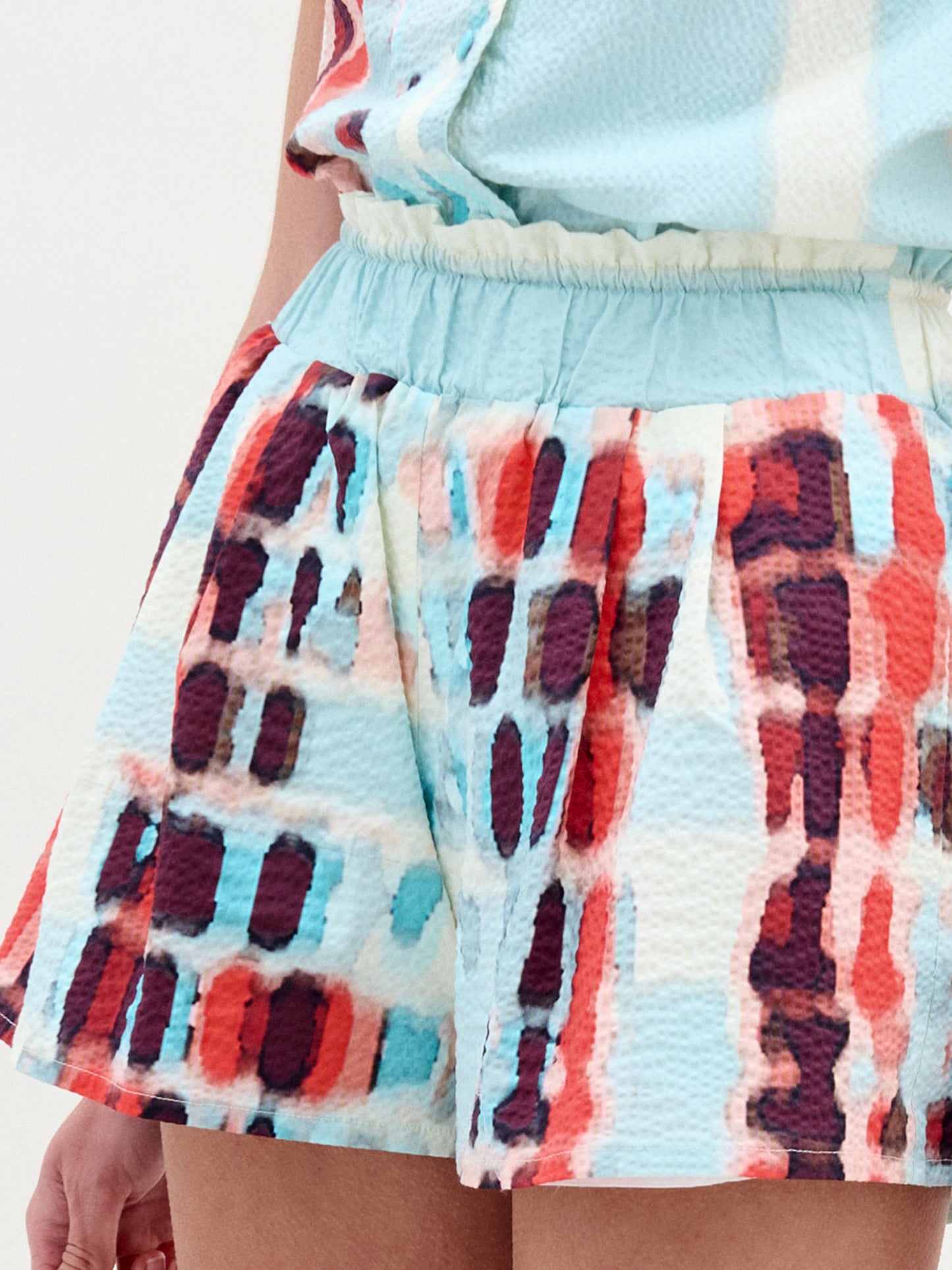 Close-up of a person wearing the Giorgio Short—high waisted, featuring a colorful red, blue, and white abstract pattern on Italian fabric—paired with a matching top. Focus is on the elastic waistband and part of the arm.