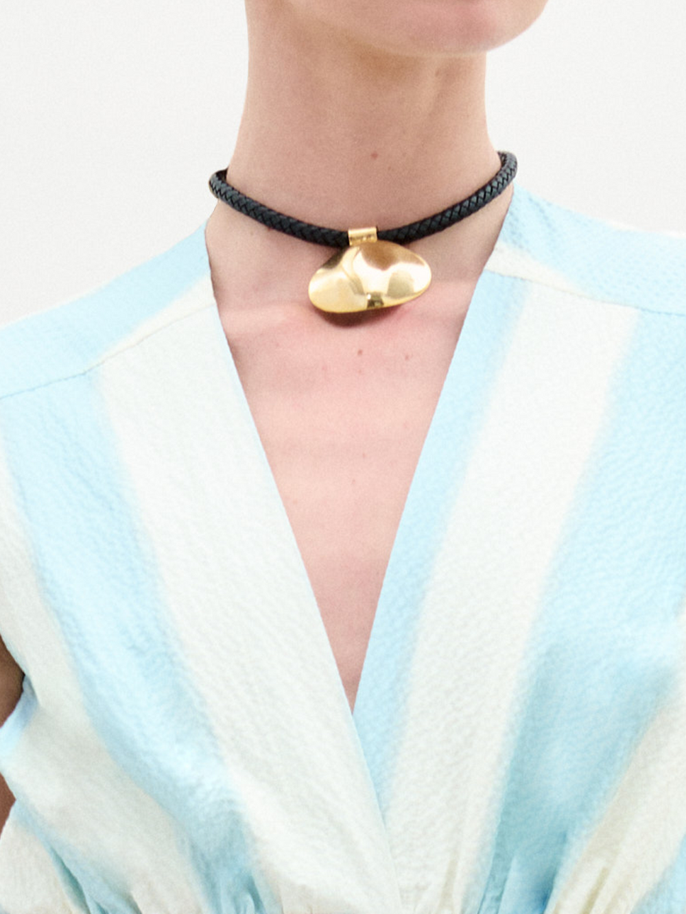 A person wears the Gimena Necklace by SVT DESIGNS, featuring a shiny gold pendant on a black leather choker. Paired with a light blue and white textured V-neck top. The image shows only the neck and upper chest. Made in Colombia.